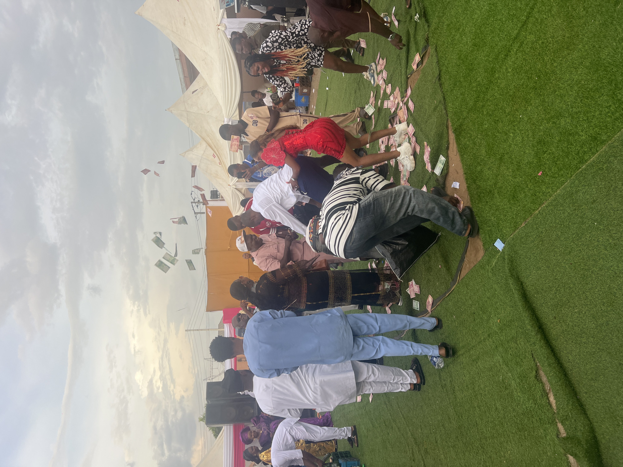 The image depicts an outdoor gathering, possibly a celebration, with numerous people. The setting appears to be on artificial turf, and there are tents and structures in the background. People are dressed in a variety of attire, with some individuals throwing money into the air. The sky is visible, with clouds suggesting either early morning or late afternoon lighting. The location is in the city of Unknown, Unknown.