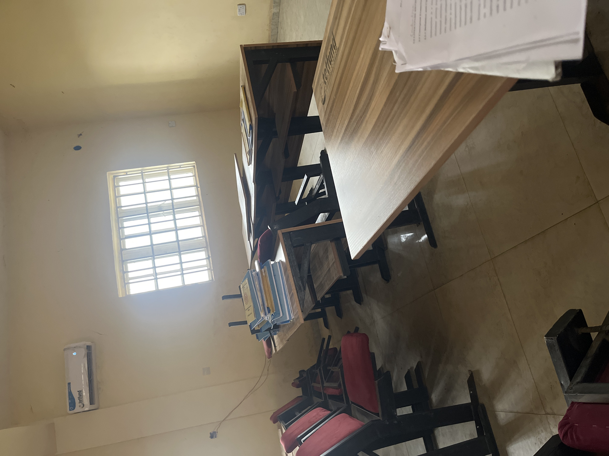 The image shows the interior of a classroom in Baganakwo, Nigeria. Several rows of wooden student desks and chairs are arranged in a tiered fashion. The desks have a light wood grain finish and are labeled "Lettlund." The chairs have red cushions. Some papers and folders are on the desks. A barred window is letting in sunlight, illuminating the room. An air conditioning unit is mounted on the wall near the window. The walls are painted in a light beige color. The floor is tiled. No people are visible in the image.