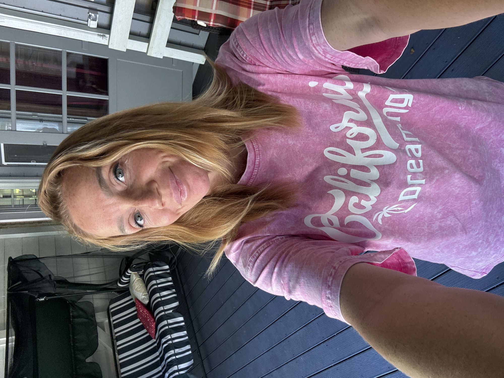 The image shows a woman with blonde hair, wearing a pink tie-dye t-shirt with white lettering that says "California Dreaming". She is smiling and taking a selfie outdoors. In the background, there is a black swing set with striped cushions, and a wooden deck. The scene appears to be a porch or a patio. There is no indication that this location is in the city of Zelenaya Roshcha, Russia.