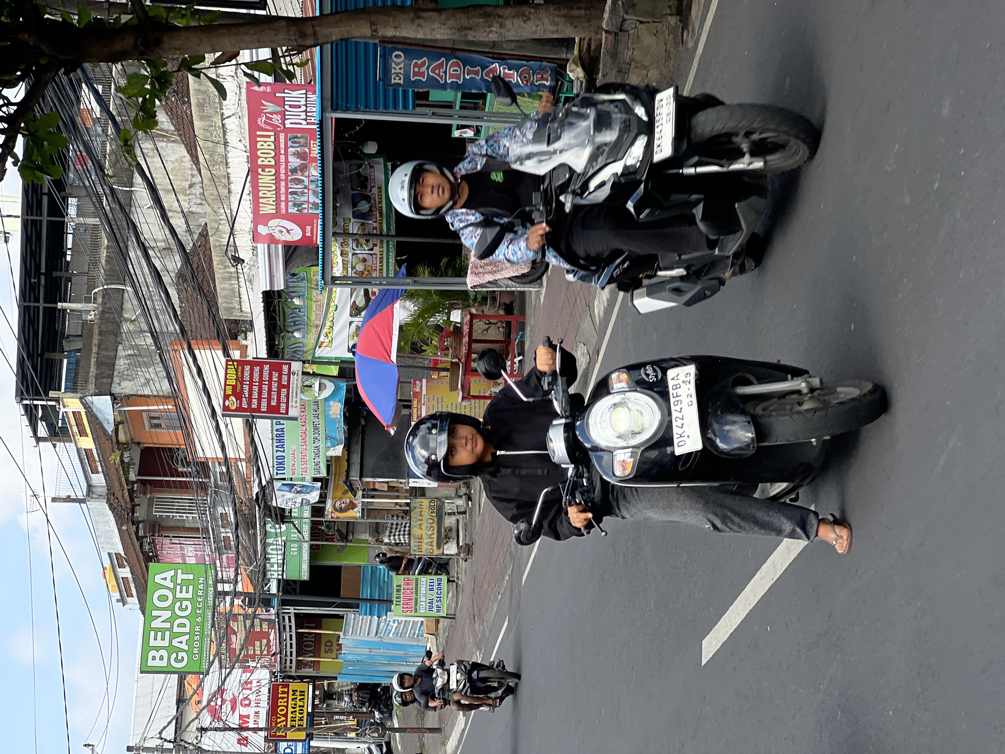 The image captures a street scene in North Kuta, Indonesia, on a cloudy day. The foreground features two individuals riding scooters on a paved road. The scooter on the right is ridden by a person wearing a black jacket with a blue and white pattern and a light blue helmet. The scooter on the left is ridden by a person wearing a black hoodie and grey pants, with a black helmet. The road has a white line marking. In the background, a bustling street is lined with various shops and businesses, indicated by numerous signs advertising goods and services. Some of the visible signs include "BENOA GADGET," "WARUNG BOBLI," and "TOKO ZAHRA P." Electrical wires crisscross overhead, common in Indonesian urban environments. The buildings are multi-story structures with varying architectural styles and colors. The overall impression is one of a lively commercial street in a Southeast Asian city.