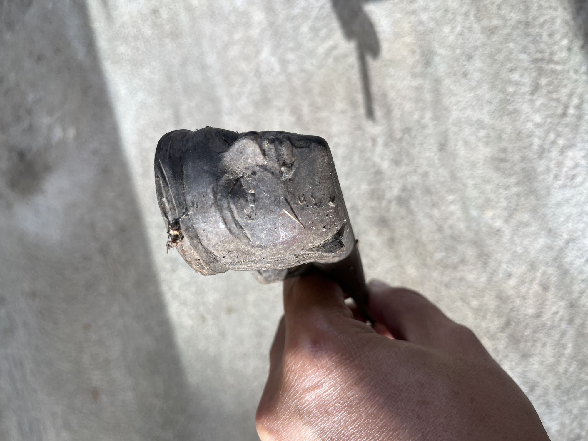 A hand holds a dark, carved wooden object with a carved face on one end. The face is detailed, with carved eyes, a nose, and lips. The object appears weathered and slightly dirty, with some debris on top of the head. The background is a textured, light-colored surface, possibly concrete, with shadows cast from an unseen light source. The setting is outdoors, under natural lighting. There is no indication of the city of Tambakreja, Indonesia within the image.