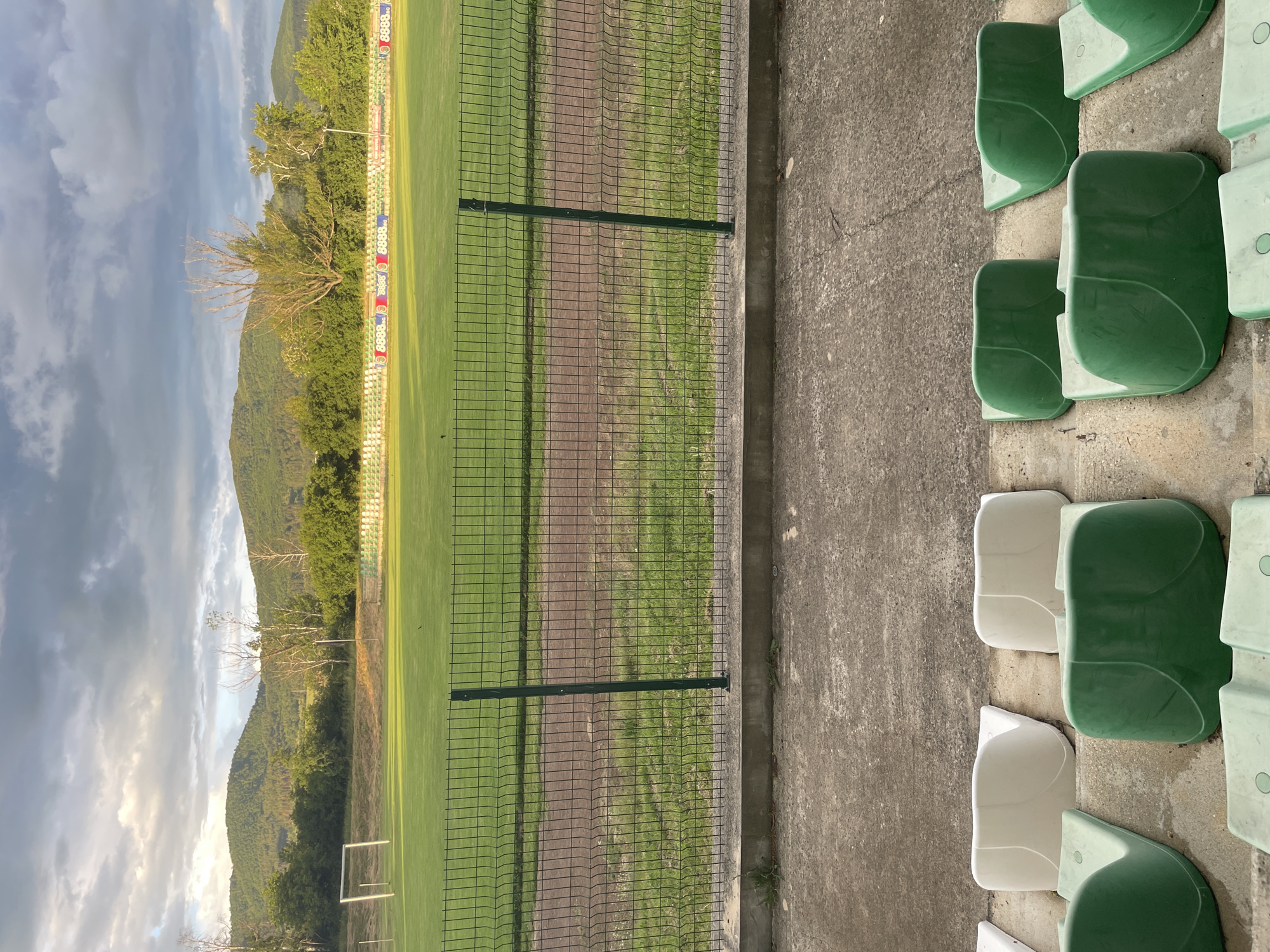 The media file captures an empty sports stadium, likely a football pitch, set against a mountainous backdrop in Ihtiman, Bulgaria. The scene is viewed from a spectator's perspective, featuring a row of green and white plastic stadium seats in the foreground, arranged on concrete steps on the right side of the frame.
A dark green metal mesh fence runs horizontally across the mid-ground, separating the seating area from the playing field. Beyond the fence, a dirt track encircles the well-maintained green grass pitch. A white goal post is visible on the far left of the field.
In the background, a line of trees and bushes, including some with prominent bare branches, forms a natural border behind the pitch. Advertising boards with the repeated text "8888" are visible along this border, likely referring to the Bulgarian betting company "8888.bg". Beyond the trees, rolling mountains covered in lush greenery stretch into the distance. The sky above is mostly overcast with soft, diffuse light, suggesting late afternoon or early evening.
No people are present, and no activity or event is taking place, giving the scene a quiet and still atmosphere. The combination of the empty stadium and the natural mountainous environment highlights the location's rural, scenic setting.