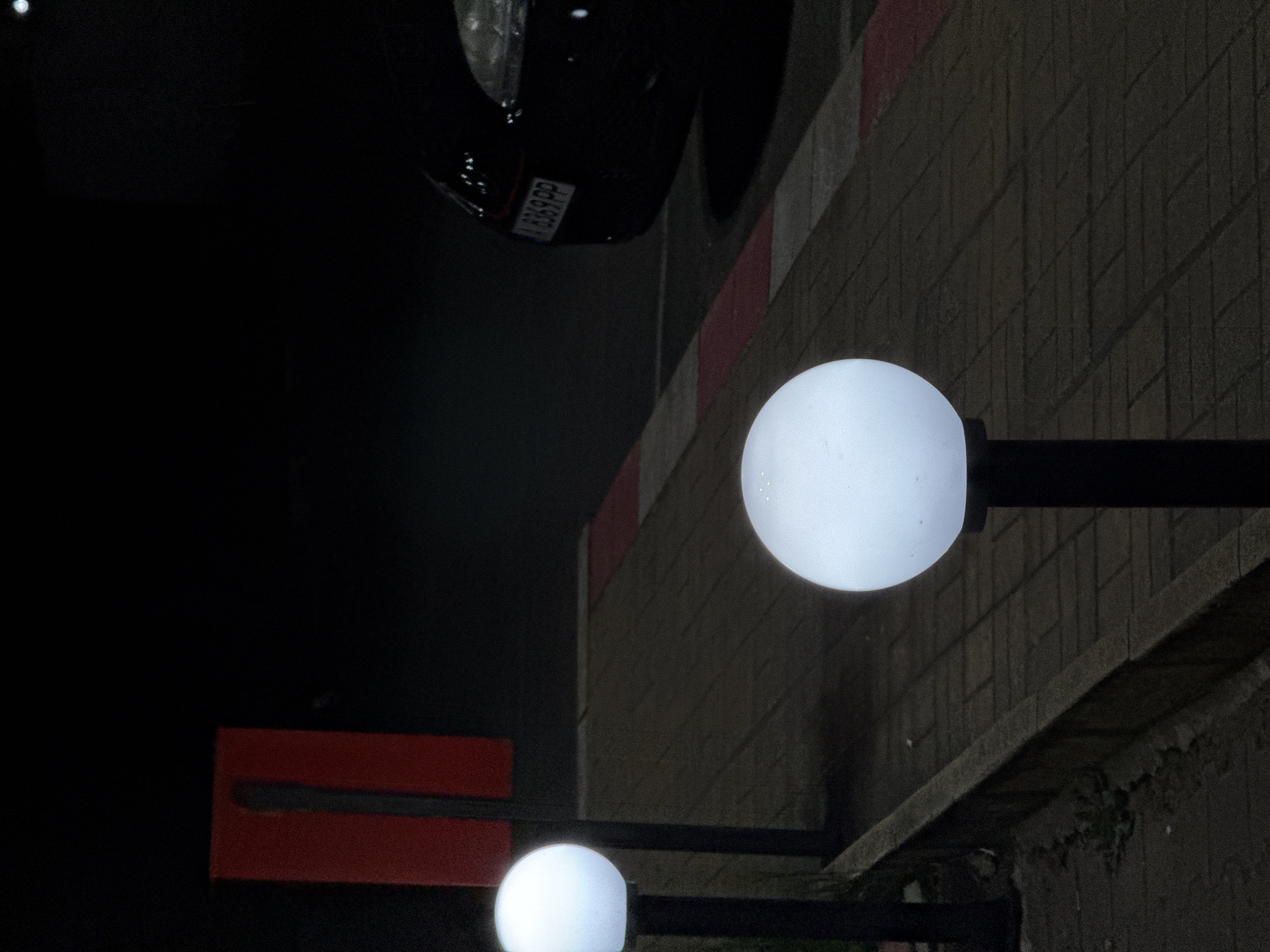 The image depicts a street scene at night in Yabalchevo, Bulgaria. Two spherical streetlights illuminate the area. A dark vehicle with license plate "B369PP" is partially visible. The streetlights are attached to black poles. A red and grey wall runs along the top of the image, and a rough stone wall is to the right. A red sign is also visible. The overall lighting is dim, with most of the scene in shadow.