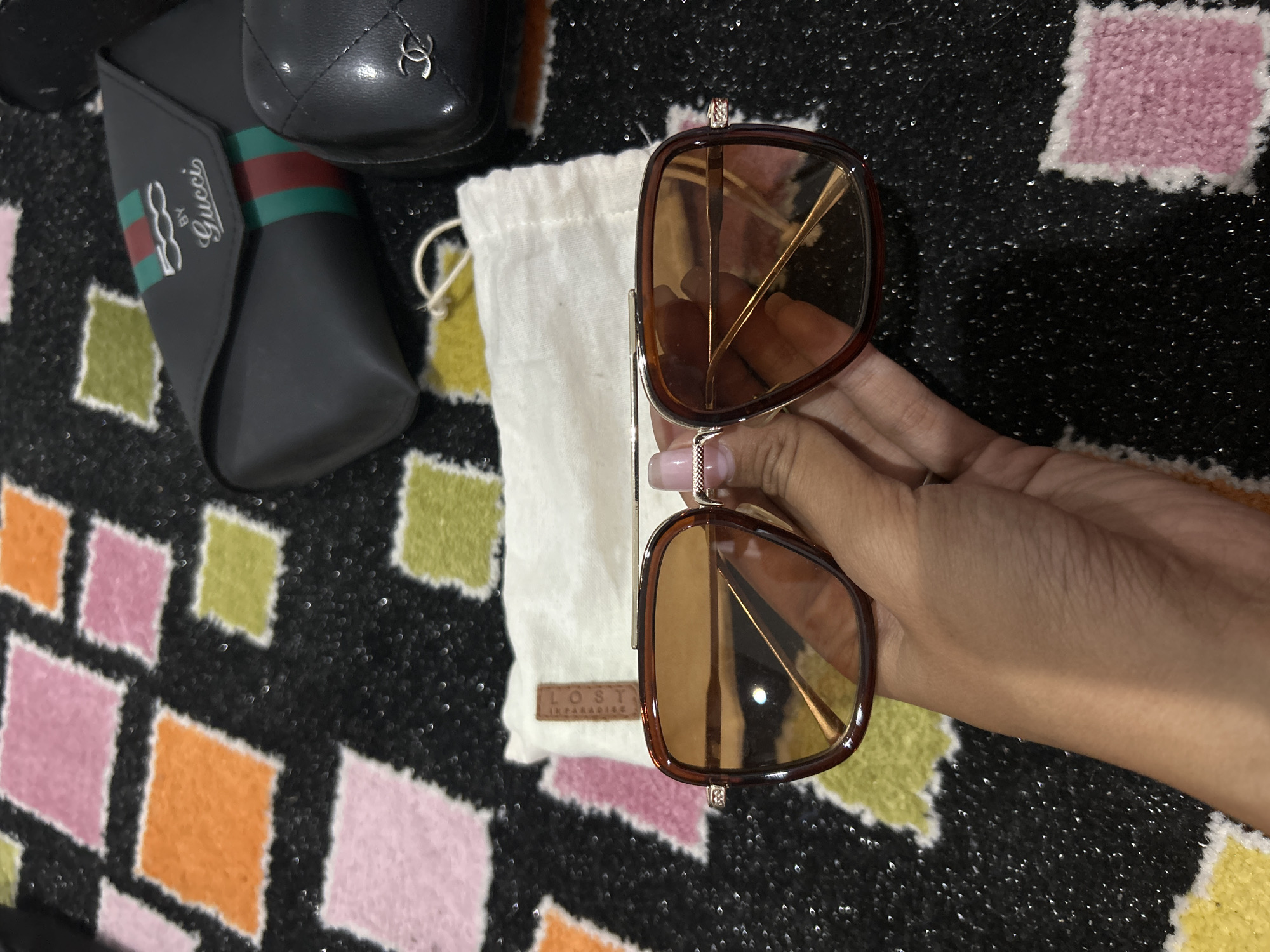 A person's hand is holding a pair of brown-tinted sunglasses with a gold frame. The sunglasses are positioned in the center of the frame, with the person's hand holding them from the bottom. In the background, there is a black carpet with a colorful diamond pattern in pink, orange, and light green. To the right of the sunglasses, a black leather sunglass case with a red and green stripe and the "Gucci" logo is visible. Next to it is a black quilted pouch with a "Chanel" logo. Behind the sunglasses, a small white cloth bag with a brown tag that says "LOST" is also visible. The image is taken from a top-down perspective. The scene is indoors, likely a home environment in Sukawati, Indonesia.