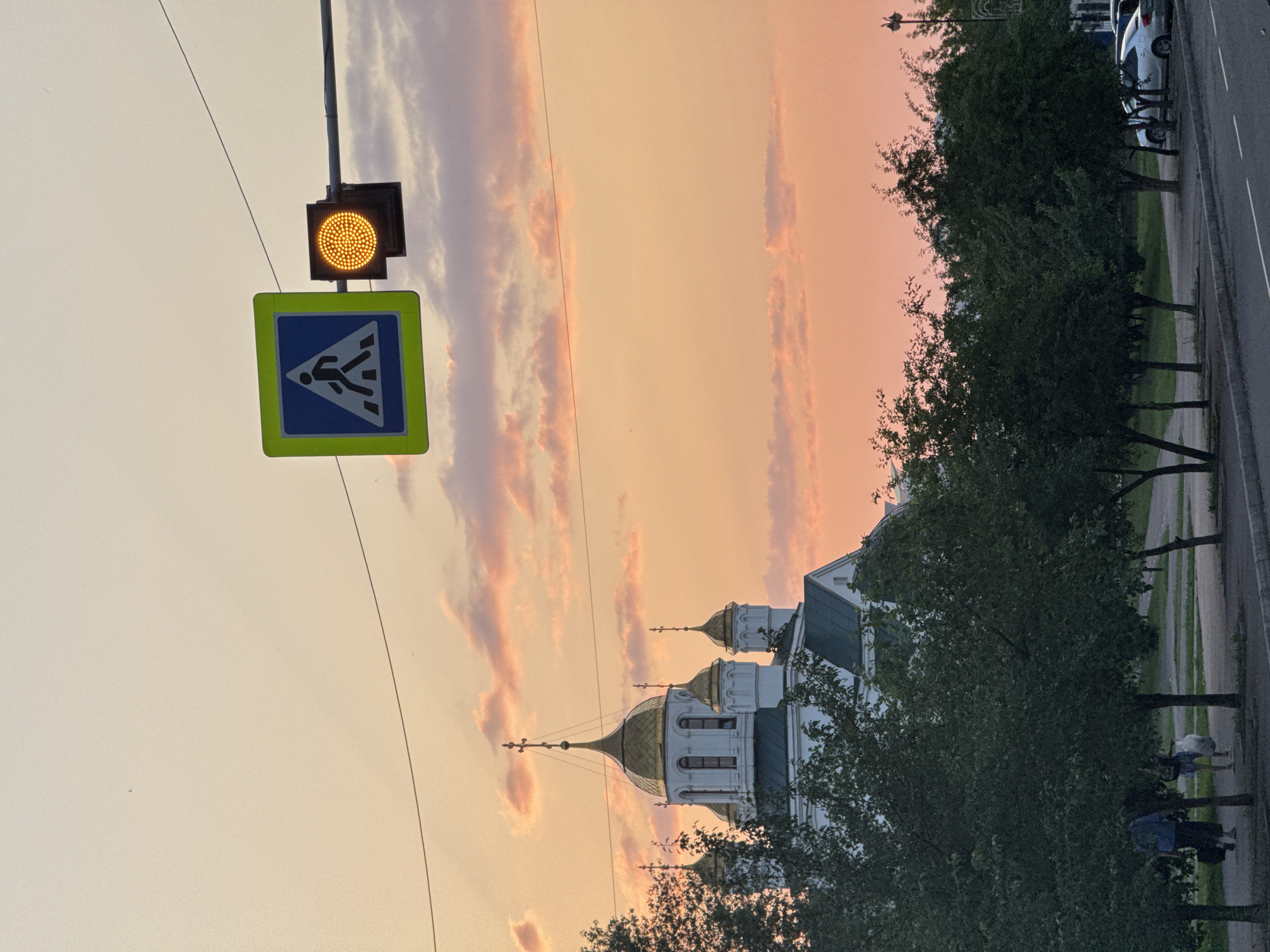 The image depicts a sunset scene in the city of Krasnoyarsk, Russia, with a pedestrian crossing sign and a yellow traffic light illuminated in the foreground. A white church with golden domes is visible in the background, partially obscured by trees. Two individuals are walking on a sidewalk beneath the trees. Cars are parked along the street. The sky is filled with soft, peachy clouds, indicating the time of day is early evening. The overall impression is one of a peaceful urban landscape at dusk.