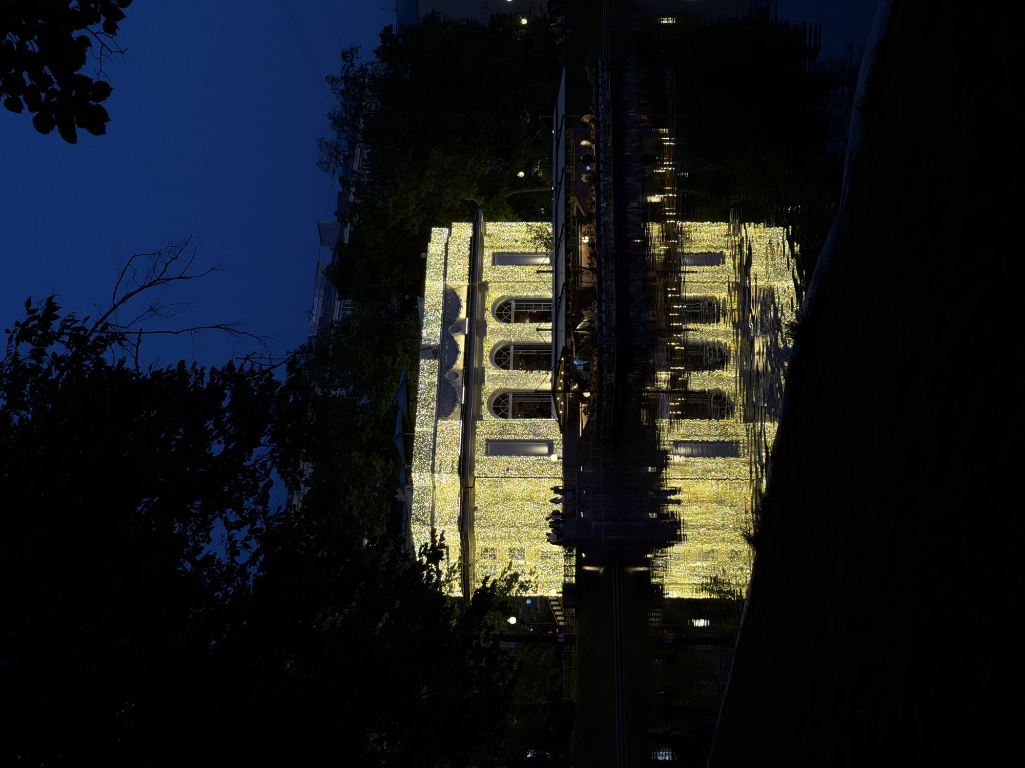 This image depicts the reflection of a building decorated with lights in a body of water at night. The building has arched windows and is covered in a dense arrangement of small, bright lights. To the left, dark foliage of trees and a view of the night sky frame the scene. The building's reflection in the water creates a symmetrical, mirror-like image. This image is likely of the GUM department store on Red Square in Moscow, Russia, during an evening lighting display.
