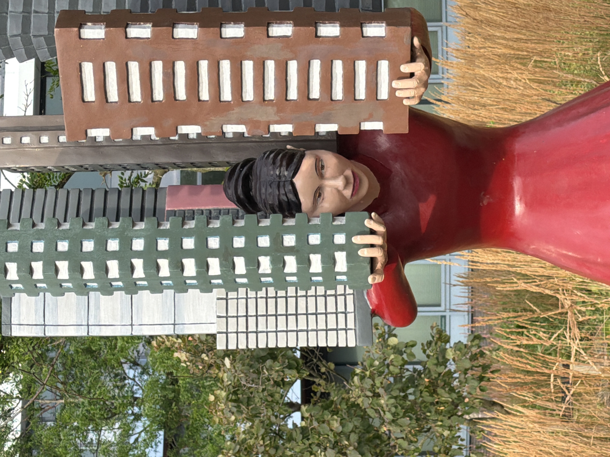 This is a photograph of a sculpture located in the city of Rijswijk, Netherlands. The sculpture depicts a person, seemingly female, wearing a red garment and holding up several representations of buildings. The buildings are stylized and vary in color and design, with some appearing as tall, rectangular blocks with rows of windows. The person's hands are visible, supporting the weight of the buildings. The sculpture is situated outdoors, with a background of greenery, dry grasses, and a building. The lighting suggests it is daytime.