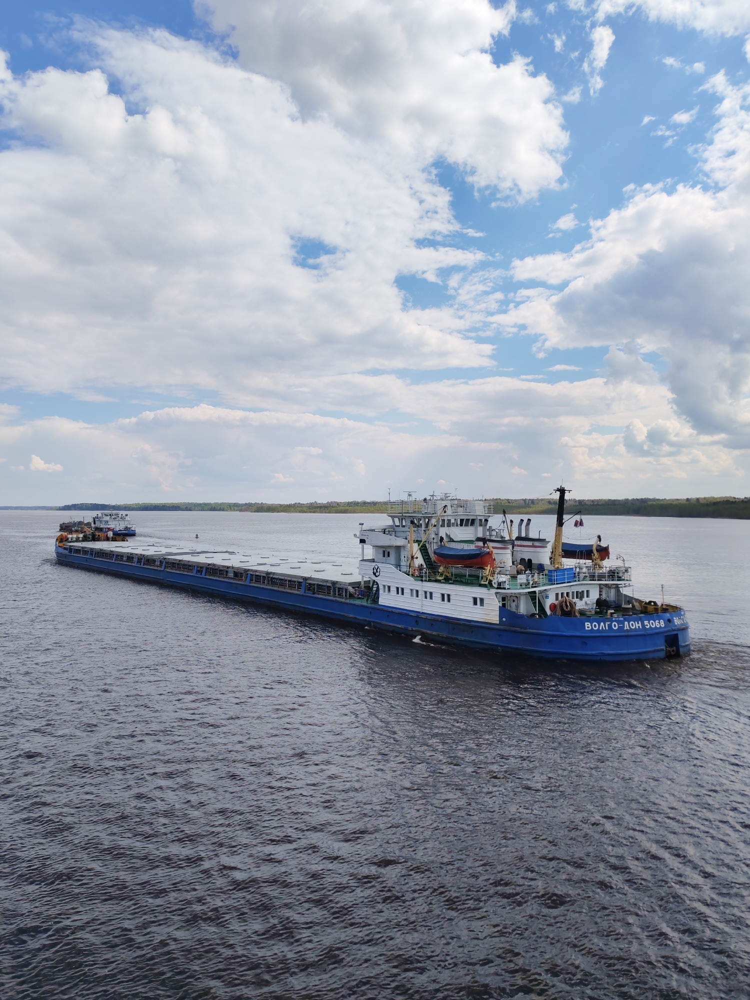 A blue and white river barge, named "ВОЛГО-ДОН 5068," is traveling on a wide body of water. The vessel consists of a flat, cargo-carrying section and a multi-level superstructure with visible lifeboats and a crane. The water's surface has small ripples. A forested shoreline is visible in the background under a sky filled with scattered white clouds. Pruzhinino is not visually indicated.