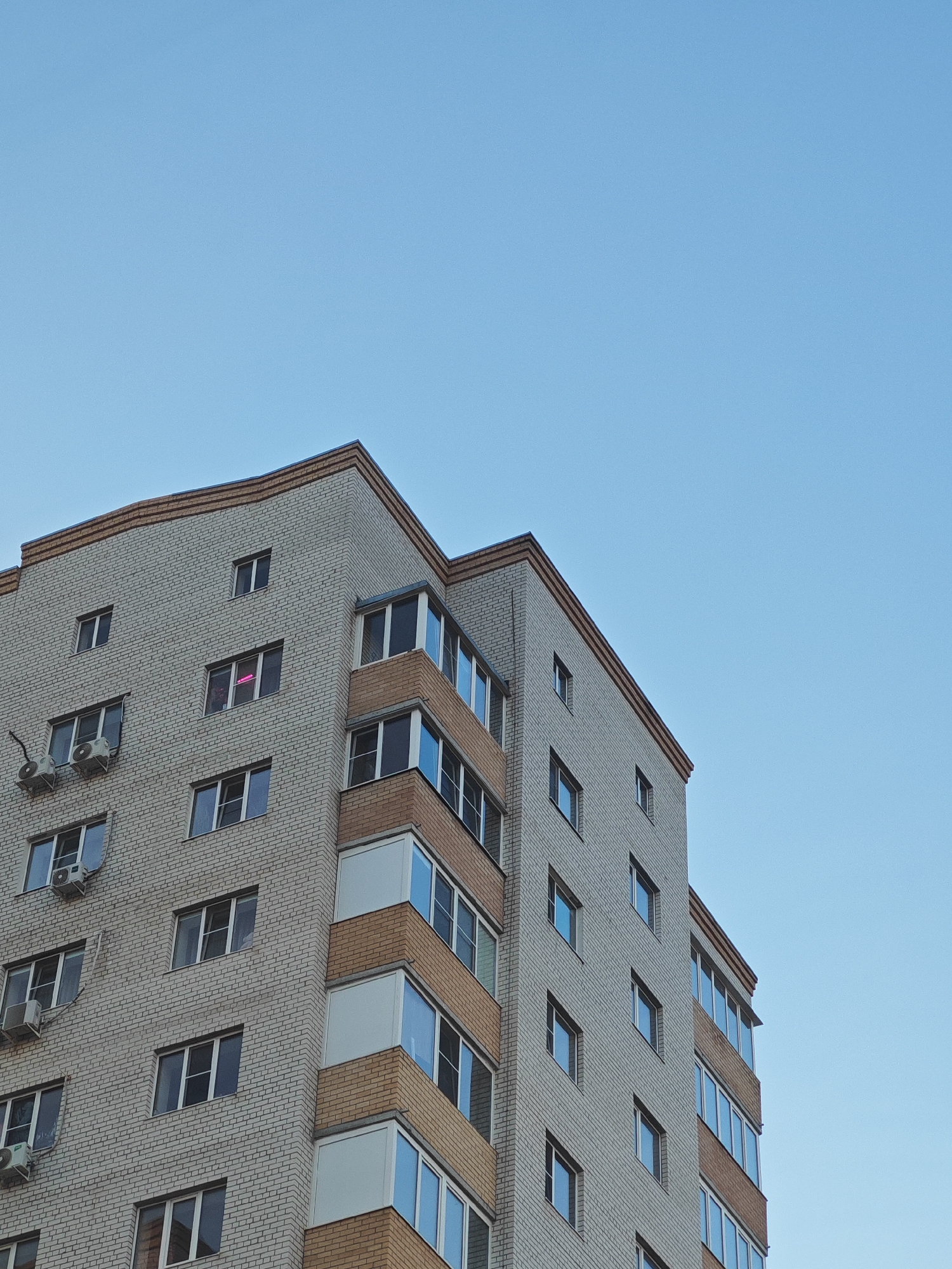 The image shows a multi-story apartment building with a light-colored brick facade and brown accents. The building has numerous windows, some of which are balconied. There are air conditioning units mounted on the exterior walls. The sky above is clear and blue. There are no people visible in the image. The building appears to be located in a city, consistent with an urban environment in Bataysk, Russia.