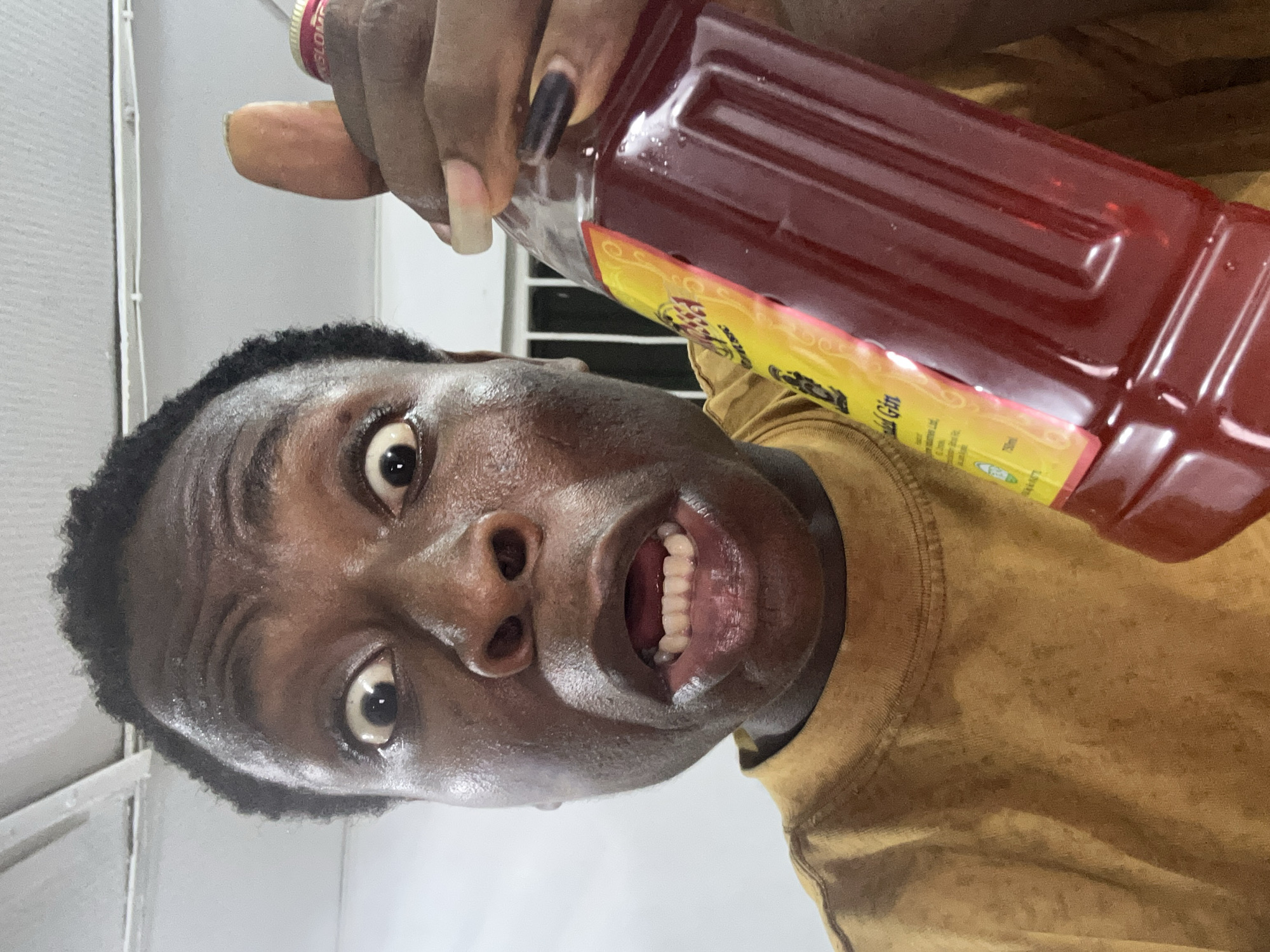 A person is holding a bottle of red liquid at an upward angle. The person has dark skin, short black hair, and is wearing a brown t-shirt. They are facing the camera with wide eyes and an open mouth, displaying their teeth. Their fingers and nails are visible, with one nail painted black. The bottle is filled with a dark red liquid and has a yellow label. The background appears to be a room with white walls and a window with horizontal blinds. The location is unknown.