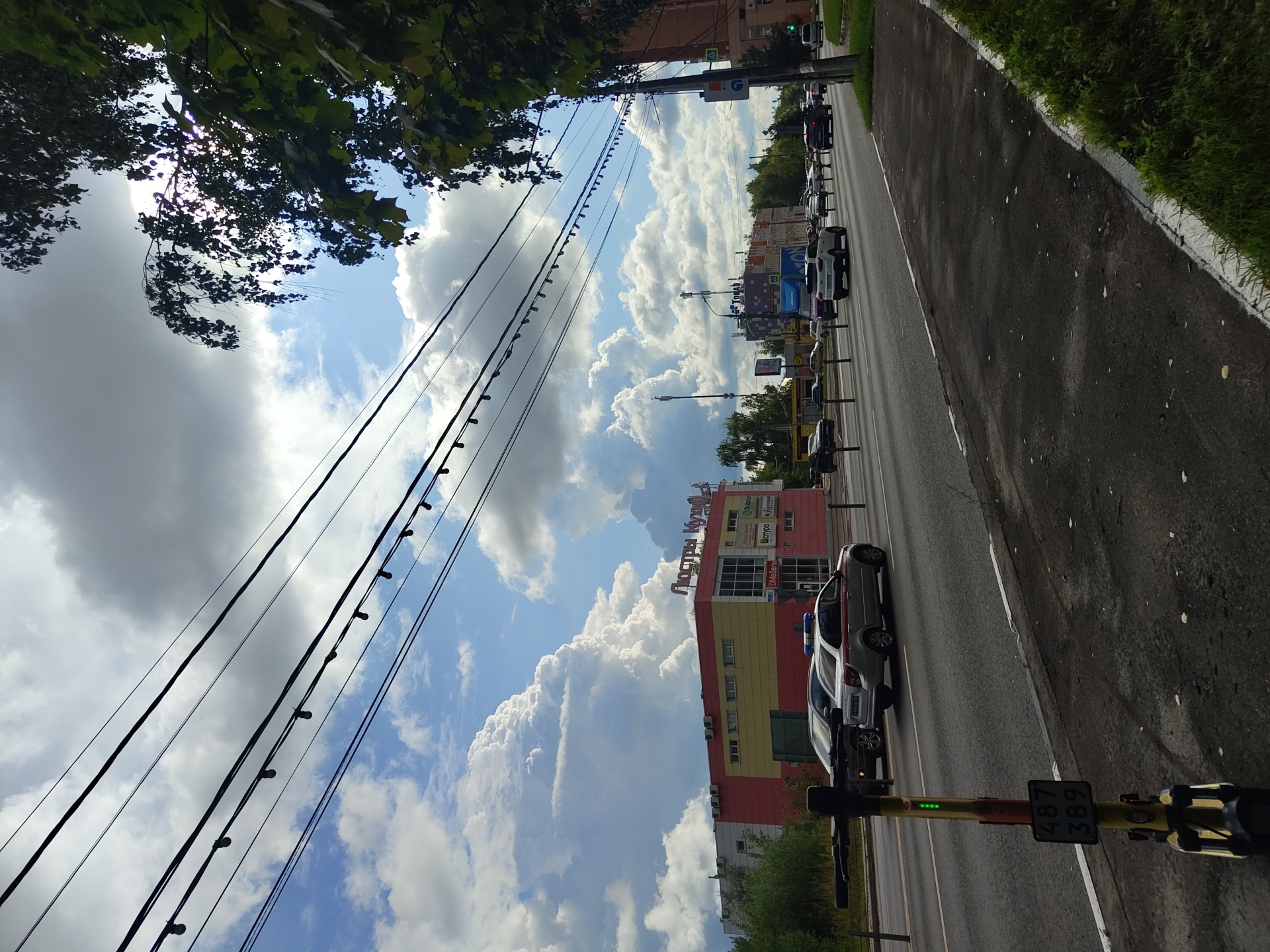 The image captures a street scene in Ramenskoye, Russia, on a partly cloudy day. The foreground features a sidewalk with green grass on the right, and a parked yellow electric scooter with license plate "487 389" and green indicator lights. A paved road with visible lane markings runs through the midground, with several cars driving in various lanes. A police car with flashing blue and red lights is visible in the right lane. On the left side of the road, a multi-story building with a prominent red and yellow facade stands, bearing the sign "Люстры Кухни" (Lustry Kukhni - Chandeliers Kitchens) in large red letters at the top. Several other buildings and trees are visible in the background. Overhead, numerous electrical wires with attached lights stretch across the frame, crisscrossing against the sky. The sky itself is filled with dramatic, large white cumulus clouds interspersed with patches of blue sky, and some darker, overcast areas suggesting approaching weather. The overall atmosphere is that of an urban street scene under dynamic weather conditions.