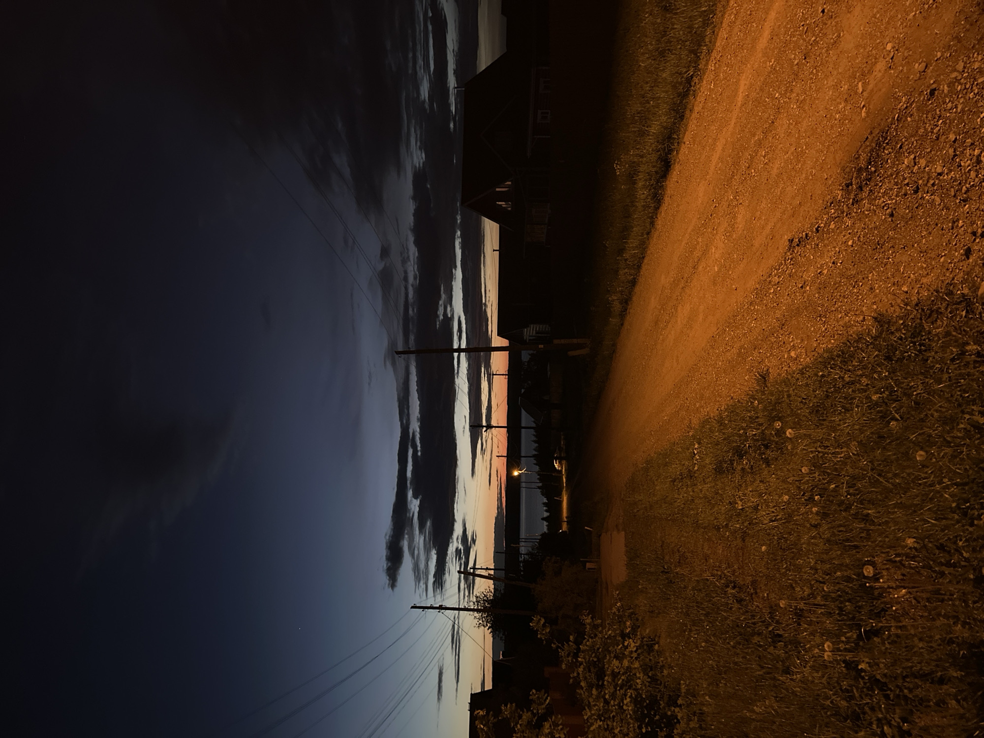 The image shows a nighttime scene in Ilyinsky, Russia. A dirt road is illuminated by a warm, amber light, bordered by grass and dandelions on the right side. On the left, the sky transitions from dark blue to streaks of orange and pink at the horizon, reflecting the fading sunset over a body of water. Silhouetted against the sky are utility poles with power lines, sparse trees, and dark outlines of buildings. The image captures a rural or suburban atmosphere under twilight.
