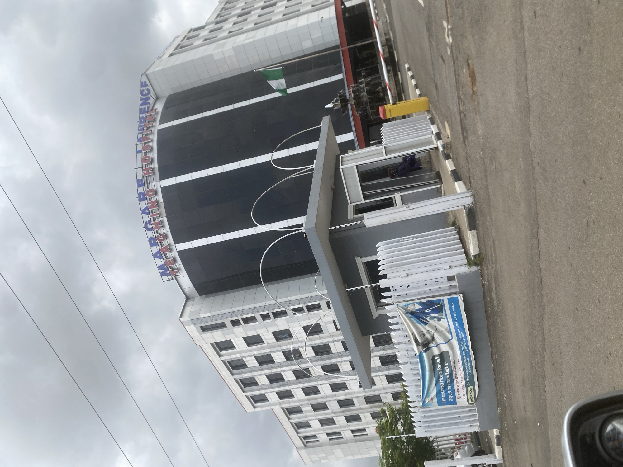 The image depicts the Marcelle Ruth Cancer Centre and Specialist Hospital in Abuja, Nigeria. The building is a modern, high-rise structure with a combination of white and black paneling. The hospital's name is vertically displayed in blue and red lettering. An entry gate is positioned in front of the building, and a banner promoting immunizations is attached to the gate. The banner indicates that immunizations for all ages are available every Wednesday. The banner includes contact information and an address of Plot 778 Akin Masokuinje Drive, Cluster 1, Riveramik Estate, Lugbe, Abuja. The Nigerian flag is visible on the building. The sky is overcast. A street is visible next to the hospital grounds.