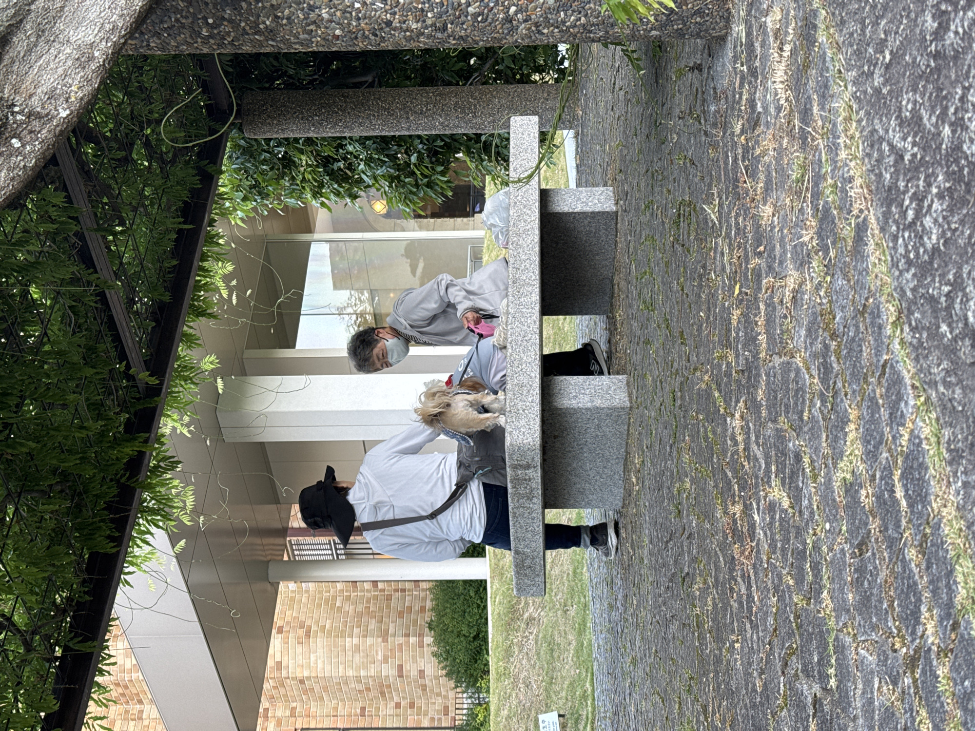Two people sit on a stone bench outdoors. One person on the left wears a white shirt, dark pants, and a wide-brimmed hat, with a cross-body bag. The other person, wearing a face mask and a light gray jacket, sits next to a small dog. They are located within the grounds of the Kyoto University. The surrounding environment includes a brick building, greenery, and a patterned stone walkway. The scene is partly shaded by an overhead structure.