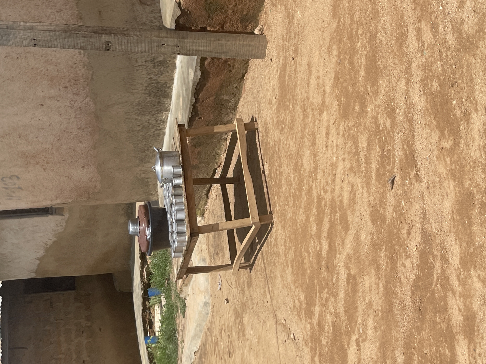 The image depicts an outdoor scene in Jos, Nigeria. The primary subject is a wooden stand holding a collection of metal containers, possibly pots or buckets, arranged in a circular pattern. A larger dark, circular object with a metal top sits below the containers. The stand is made of rough-hewn wood, suggesting a rustic or makeshift construction.
The setting appears to be an unpaved area with dry, sandy soil. In the background, there is a building constructed with concrete or mud walls. Some greenery is visible near the bottom right of the frame, hinting at vegetation. The light suggests it is daytime, and the overall atmosphere seems arid, consistent with a dry climate. No people are visible in the image.
There is some faint text visible on the wall in the upper left corner of the image, appearing as a few blurred characters. The image is oriented vertically, and due to the rotation, the ground appears to be the sky and the sky the ground. There are no specific indicators of the time of day beyond it being daylight. The weather appears to be dry and sunny. The scene conveys a sense of everyday life, likely related to storage or preparation of goods.