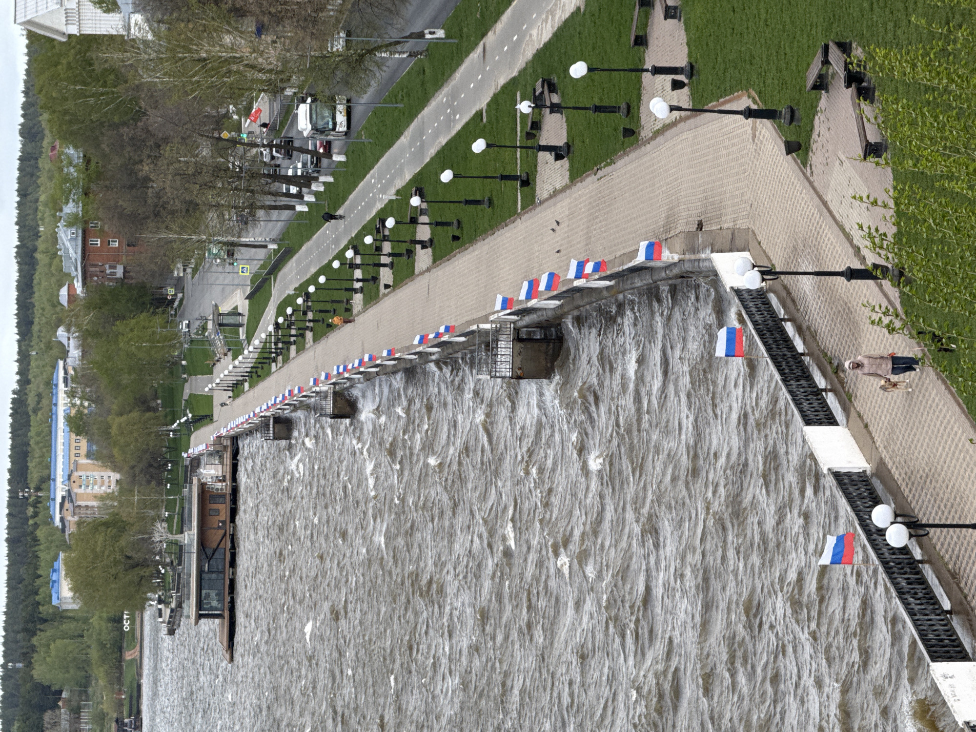 The image shows a section of the Votkinsk, Russia embankment on a body of water. The water is rough, indicating a high water level or strong current. A paved promenade runs along the water's edge, lined with black lamp posts with white globes. The embankment wall is adorned with small Russian flags. A road runs parallel to the promenade, with parked vehicles visible. Buildings and trees are visible in the background, indicating an urban setting. A single person is present on the promenade near the end of the paved section. A dock juts out into the water.