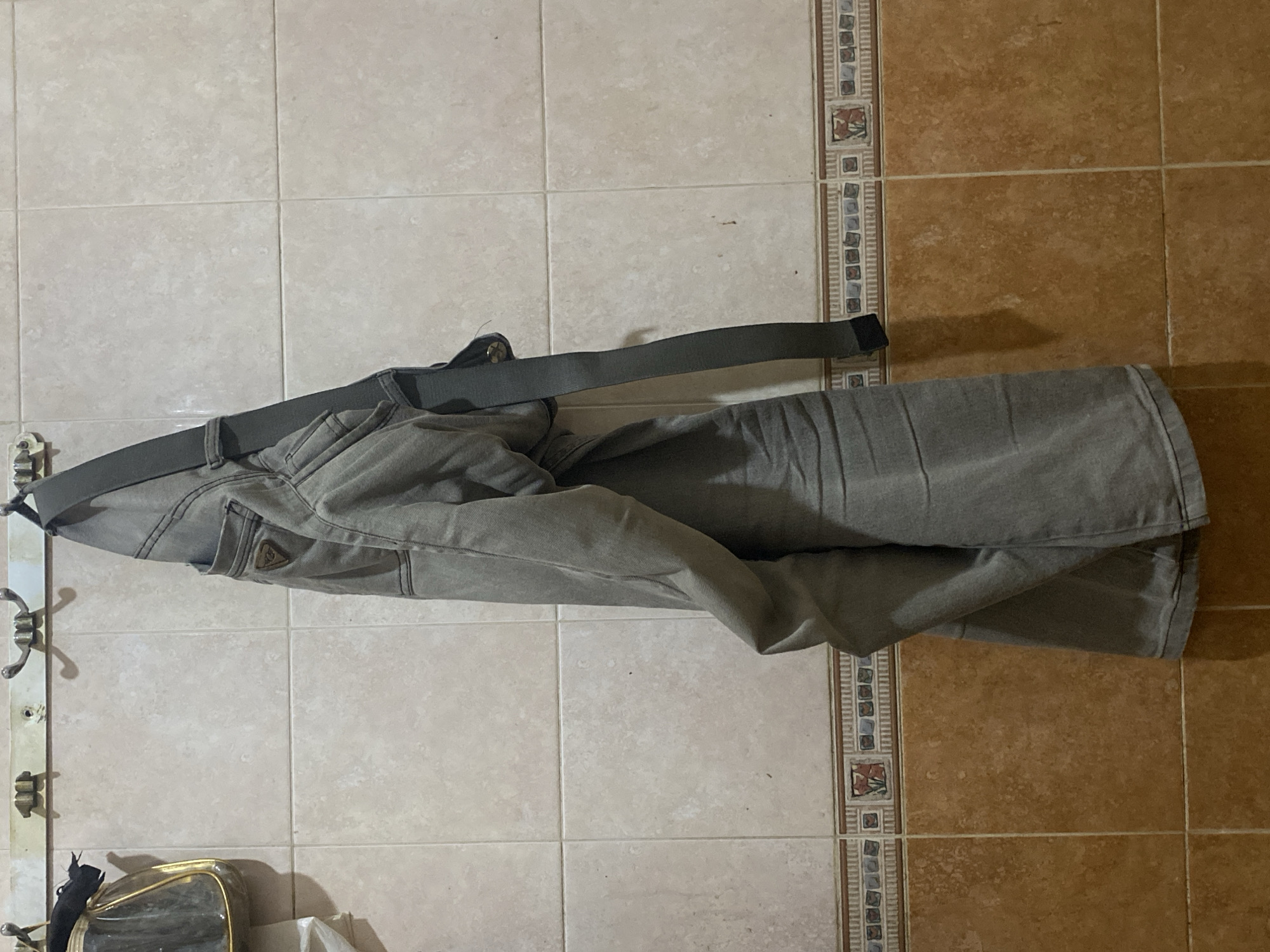 A pair of gray pants is hanging from hooks on a tiled wall in a bathroom. The pants have a dark gray belt attached. To the left of the pants, a small, clear plastic and gold handbag is also hanging from a hook. The wall is tiled with light beige squares and has a decorative border of smaller tiles with an intricate pattern, separating it from the lower section of the wall, which is tiled with larger, terra cotta-colored square tiles. The scene is likely in a bathroom in a residential building in North Jakarta, Indonesia, given the typical tile patterns and hook styles found in the area.