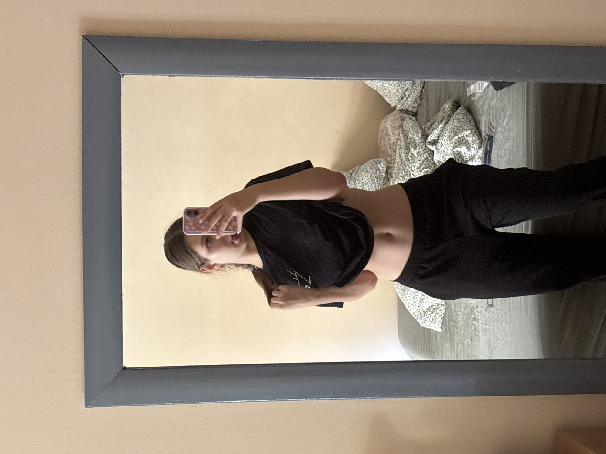 A reflection is visible in a rectangular mirror with a dark gray frame, which is mounted on a beige wall. Reflected in the mirror is a young woman holding a smartphone in her left hand to take a selfie. She is wearing a black t-shirt that is partially raised to expose her midriff, and black sweatpants. Behind her is a bed with a patterned comforter in shades of green and white. The location, Bečići, Montenegro, is not ascertainable from the visual data.
