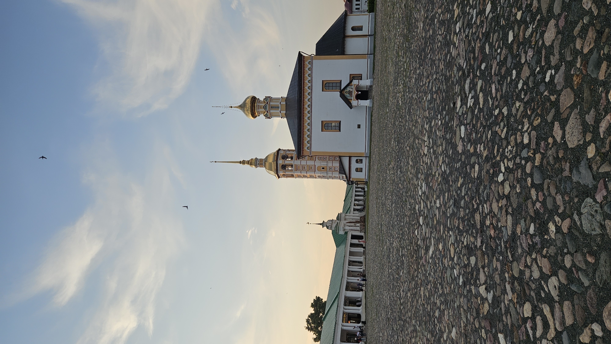 The image shows a wide cobblestone plaza in front of two churches and a long, colonnaded building in Suzdal, Russia. The church on the right is white with golden domes and a bell tower. The church on the left is partially obscured by the colonnaded building but appears to have a single dome. The colonnaded building is green and white, with numerous columns supporting a green roof. Several people are visible walking in the plaza and near the colonnaded building. The sky is blue with scattered white clouds, and there are several birds flying in the sky. The cobblestones are varied in size and color, and the scene appears to be set in the late afternoon or early evening, with warm light illuminating the buildings.