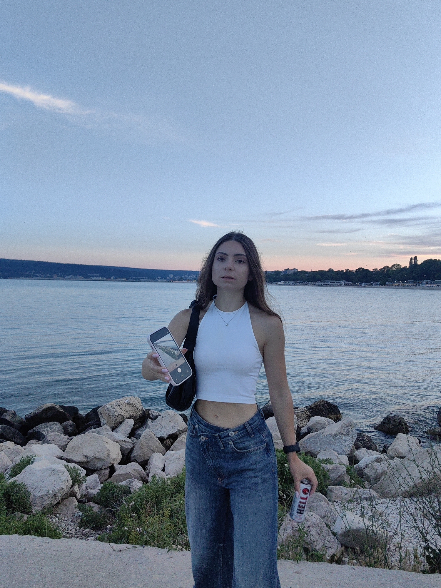 A young woman stands on a rocky shore at dusk, holding a smartphone in her left hand, displaying a reflection of the scenery. She is wearing a white crop top and blue denim jeans. In her right hand, she holds a can of beverage. The background features a calm body of water, with a distant shoreline and a hint of a city skyline. The sky above is a gradient of blue and pink, indicating the transition from day to night. The overall scene is peaceful and serene.