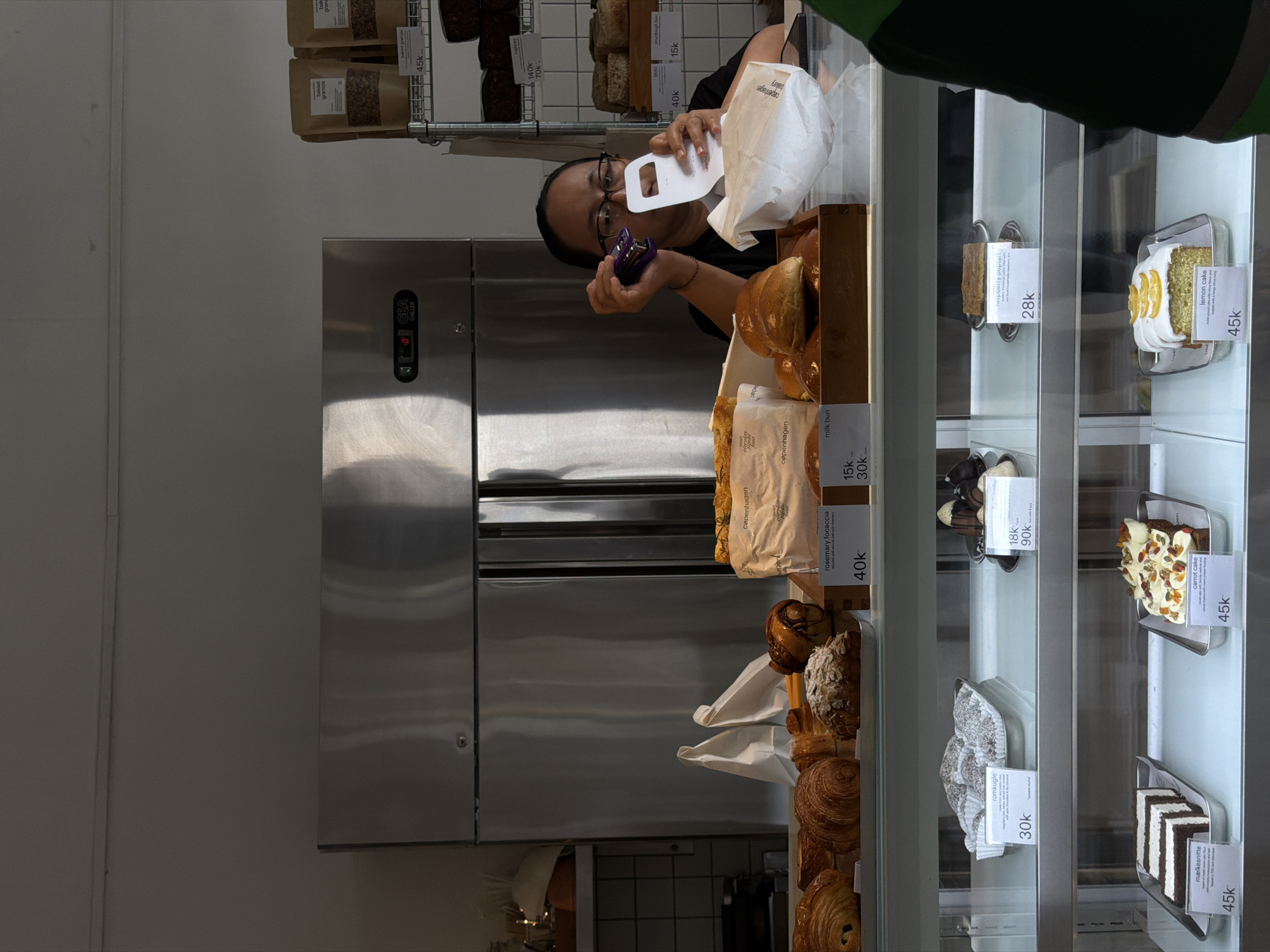 The image shows a woman with glasses and her hair pulled back, wearing a black t-shirt, standing behind a counter in what appears to be a bakery. She is holding a purple stapler in her right hand and a white paper bag in her left hand, and she is looking at the stapler. In front of her is a display case filled with various baked goods, including croissants, buns, and cakes. The display case is illuminated, and the baked goods are neatly arranged on trays. Behind the counter, there is a large stainless steel refrigerator. The environment appears to be a retail setting. Based on the context provided, this scene is located in Mengwi, Indonesia.