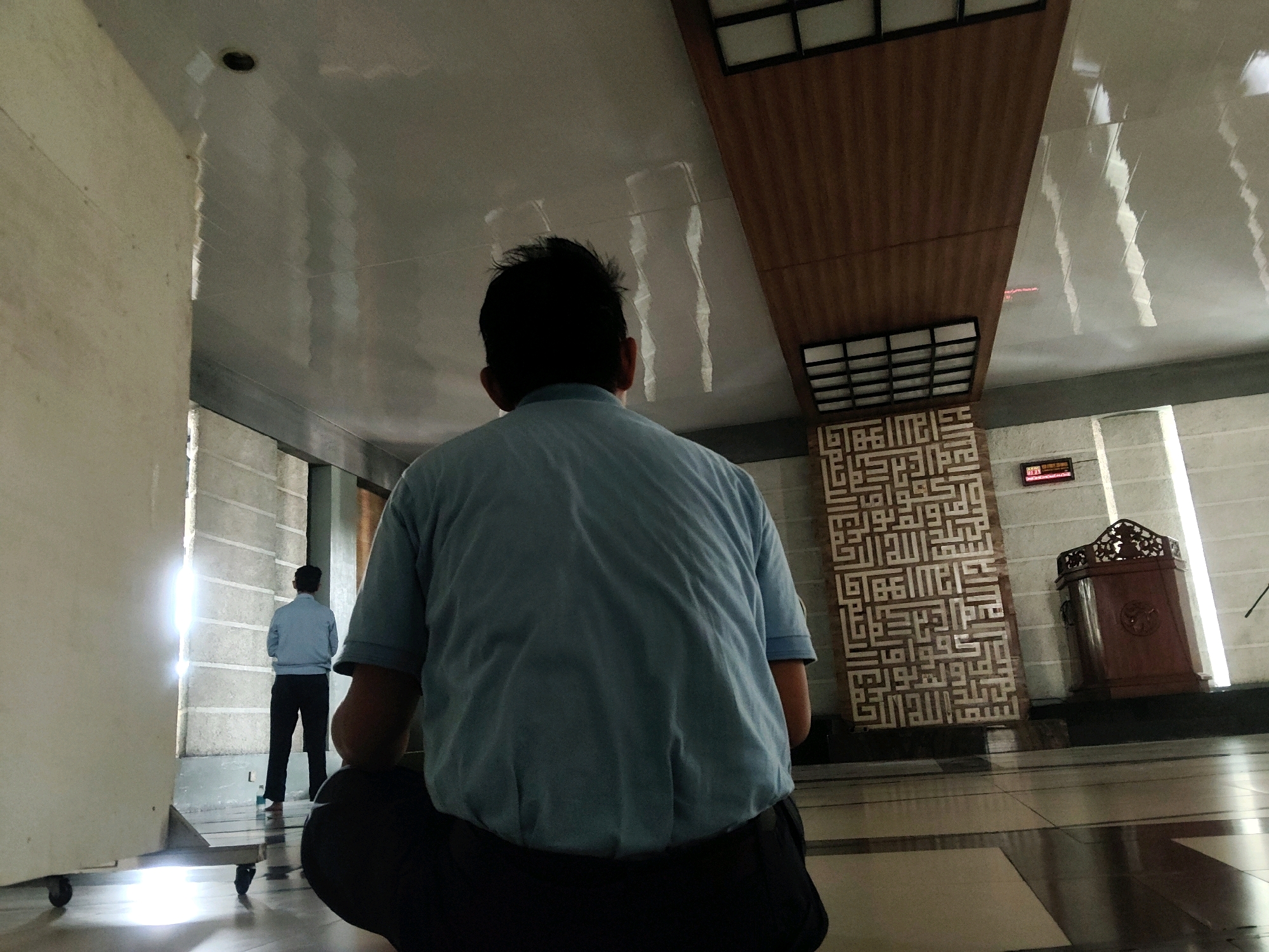 In an interior space with a reflective ceiling and tiled floor, a person in a light blue short-sleeved shirt and dark pants is seated on the floor, facing away from the camera. To the left, a large, off-white structure with wheels is partially visible. In the background, another person in a light blue long-sleeved shirt and dark pants stands facing a brightly lit wall with vertical textured lines. Further in the background, on the right, a decorative wooden structure with intricate carvings is positioned next to a vertical surface adorned with geometric Arabic calligraphy. A digital clock displaying "11:04" is mounted on the wall between the calligraphy and the wooden structure. The overall lighting suggests it is daytime. The scene appears to be a public space, possibly a prayer hall or a communal area within a building in Mekarmukti, Indonesia.