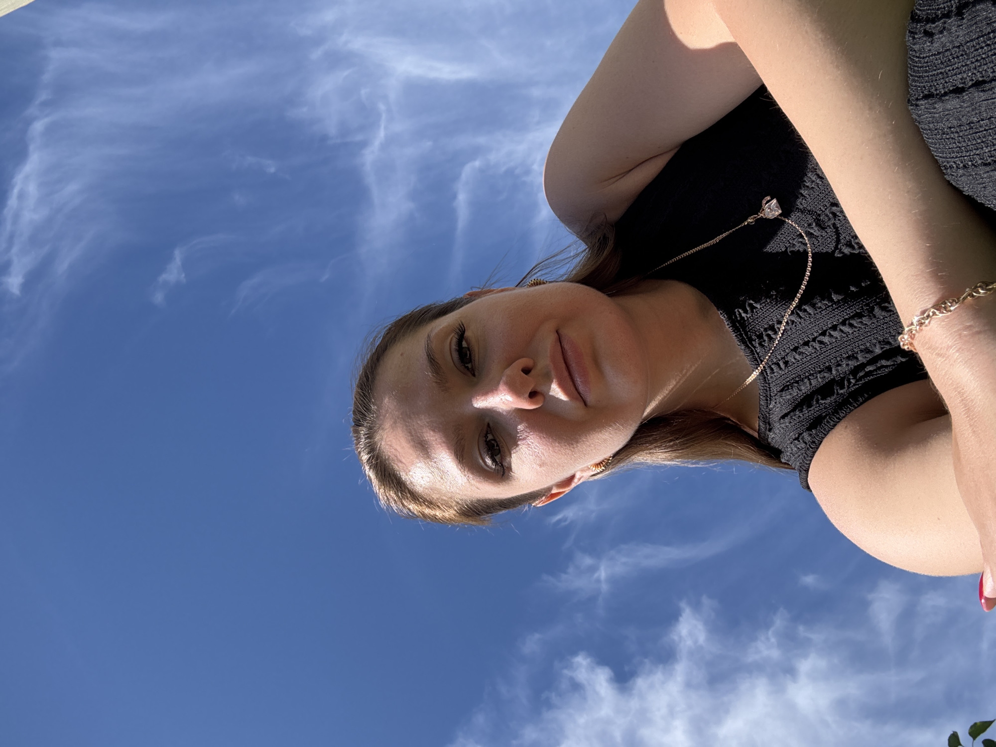 A low-angle, waist-up shot shows a young woman with light brown hair wearing a black top and gold jewelry, including a necklace with a small pendant and a bracelet. She is looking at the camera with her head tilted slightly. Her makeup includes winged eyeliner. Her left arm is bent and resting across her chest. The background is a clear blue sky with wispy white clouds. The lighting suggests it is daytime. There are no discernible city landmarks or environmental features to indicate a specific location.