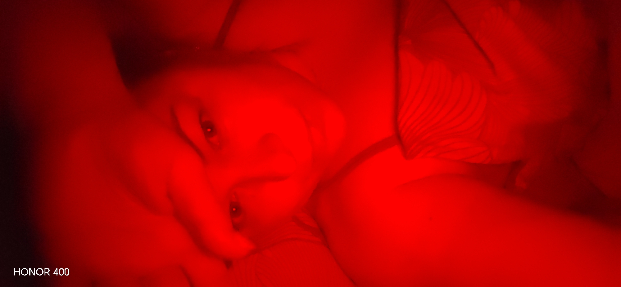 The image captures a close-up of a person, likely female, lying down in a low-light setting dominated by a vivid red hue. The entire scene is drenched in red light, creating a dramatic and somewhat mysterious atmosphere, suggesting the use of colored artificial lighting. The subject's face is partially visible and turned towards the viewer, with their eyes distinctly discernible, showing bright reflections in the pupils, indicating a light source. The features are rendered with a soft focus, giving a slightly blurred, dreamlike quality, although the gaze appears direct. On the right side of the frame, a striped pattern is visible, likely belonging to clothing or bedding. The intimate framing and artificial red lighting suggest an indoor setting, possibly a bedroom, during nighttime or in a darkened room. In the bottom left corner of the image, the text "HONOR 400" is visible.
