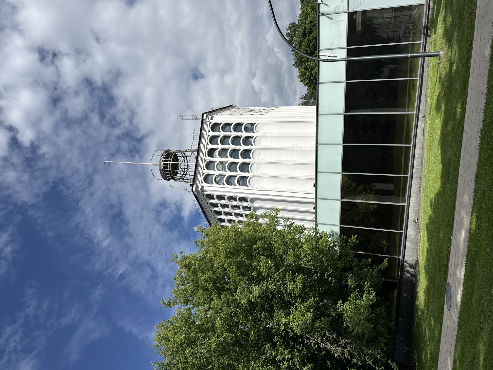 This is a vertical, eye-level, outdoor photograph of a modern building with distinctive architectural features, set against a bright blue sky with scattered white clouds. The building is white and has a tall, slender tower-like section adorned with rows of semi-circular windows. The tower is capped with a metal structure. Adjacent to this section is a modern extension with a facade of large, dark, reflective glass windows, set in a light grey frame.
To the left and front of the building, a large, lush green tree with dense foliage partially obscures the lower part of the structure. A neatly maintained lawn runs alongside the building, bordered by a paved walkway. The lighting suggests it is daytime, with sunlight casting shadows on the grass. The overall impression is one of contemporary architecture set within a well-kept urban or campus environment. The image does not depict any people or specific events. There is no visible text in the image.