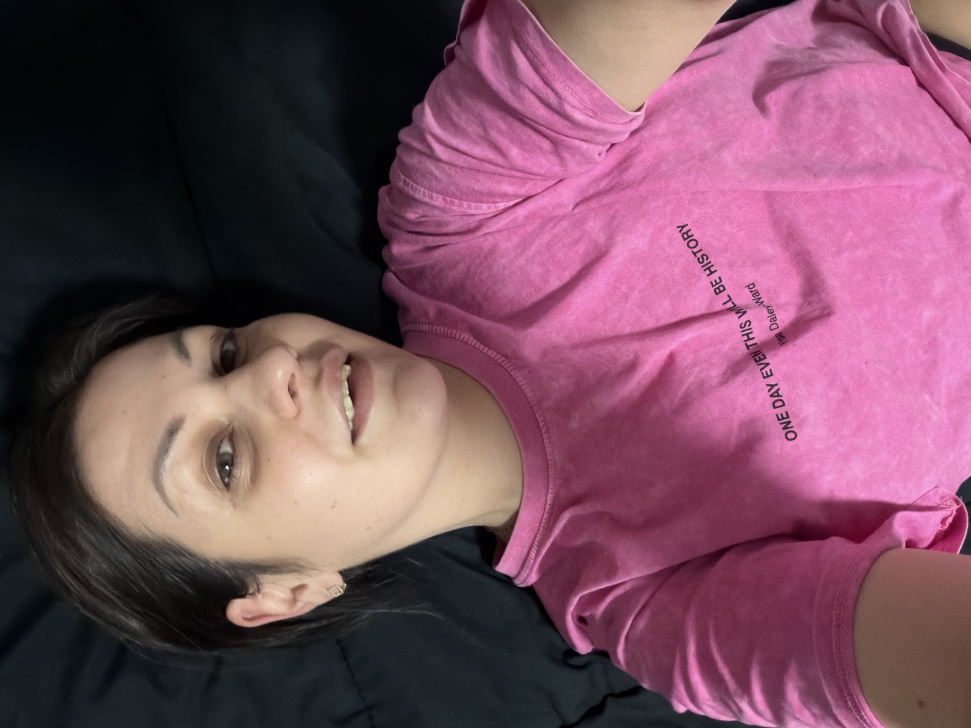 The image is a close-up, overhead shot of a young woman lying on her side on a dark surface, likely a bed. She is wearing a bright pink t-shirt with black text printed on it that reads "ONE DAY EVEN THIS WILL BE HISTORY" followed by "Yrsa Daley-Ward" in smaller print. Her hair is dark brown and styled short, framing her face. She has light skin and is looking directly at the camera with a slight smile. Her mouth is slightly open, revealing her teeth. She is wearing a small, gold earring in her left ear. The background is a dark, textured surface, likely bedding, which is out of focus. There are no discernible landmarks or objects that would place the image in Sochi, Russia.
