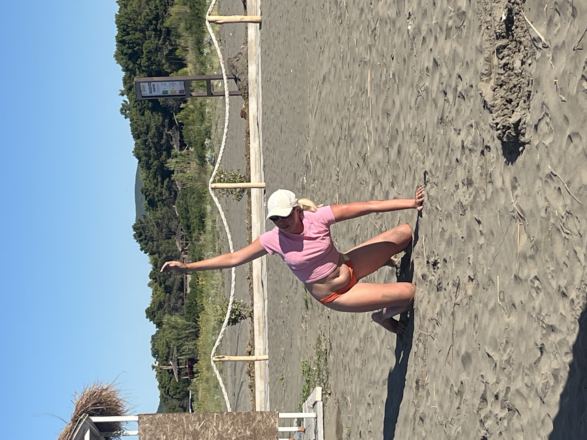 A young woman with blonde hair, wearing a pink t-shirt and orange bikini bottoms, kneels on a sandy beach. She is wearing a white baseball cap and her right arm is extended upwards. The beach is lined with a rope fence and in the background there is a line of trees and a clear blue sky. A small beach hut with a thatched roof is visible on the left side of the image. The scene appears to be in a natural outdoor setting, possibly a resort area, given the amenities visible. The woman's pose suggests she may be exercising or stretching. The sand is a dark grey color and appears somewhat wet or damp.