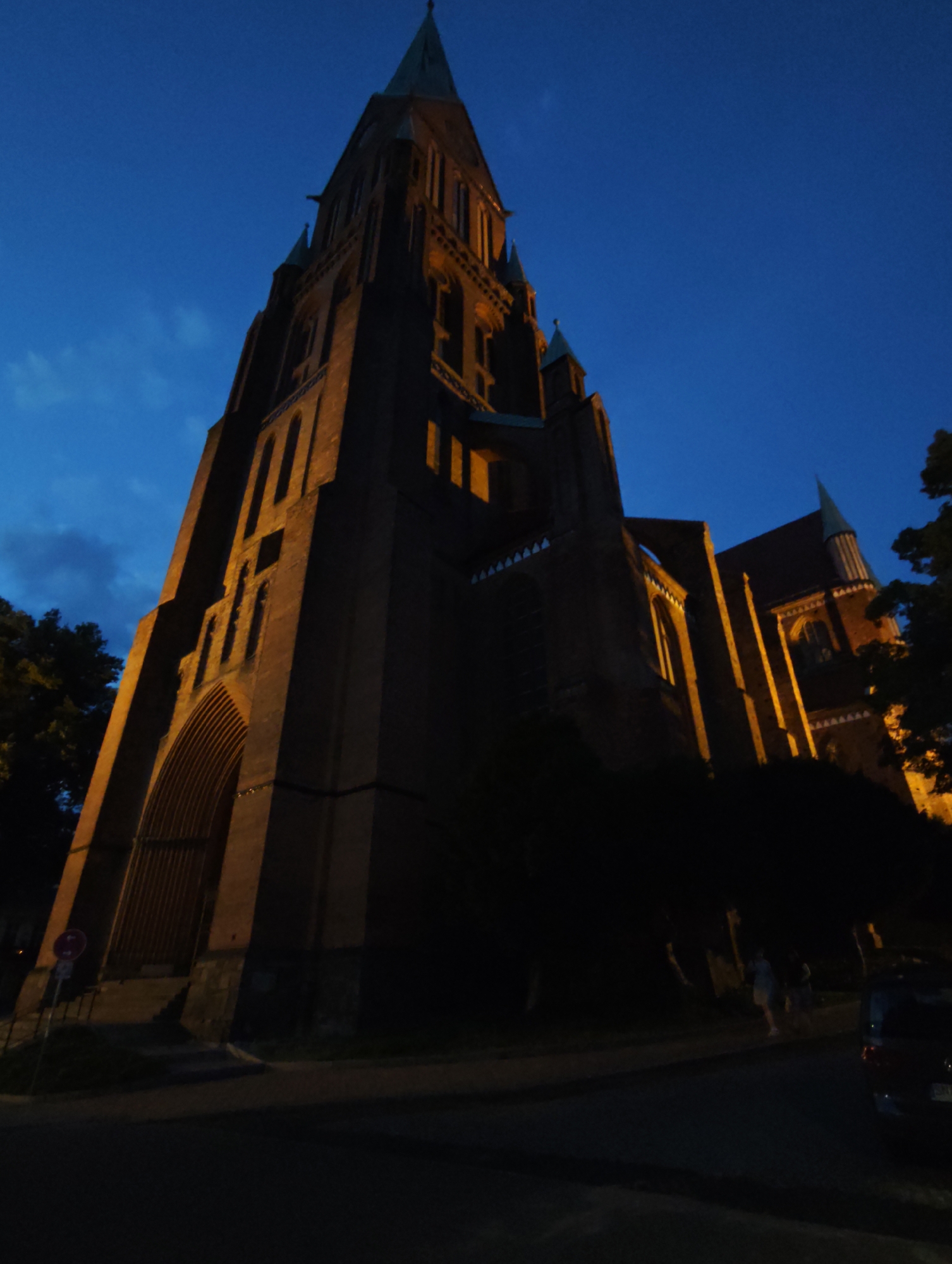 The image shows a low-angle, wide shot of a large brick church in Schwerin, Germany, at dusk. The sky is a deep blue with some scattered clouds. The church is illuminated by warm, artificial lights, highlighting its gothic architectural features, including tall spires, arched windows, and intricate stonework. The facade of the church is predominantly brick, with some stone accents. The lighting casts long shadows, emphasizing the scale and grandeur of the building.
In the foreground, a paved road or sidewalk runs in front of the church. To the left, there is a partial view of a wrought-iron gate or fence leading up to the church entrance. A red and white circular sign is visible near the gate. Several trees with dark foliage are scattered around the base of the church and along the road.
On the right side of the road, two individuals are walking away from the camera. They are indistinct due to the low light conditions and distance, but appear to be casually dressed. Further to the right, the rear of a dark-colored vehicle is visible, parked on the side of the road. The overall atmosphere is one of quiet evening serenity in an urban setting.