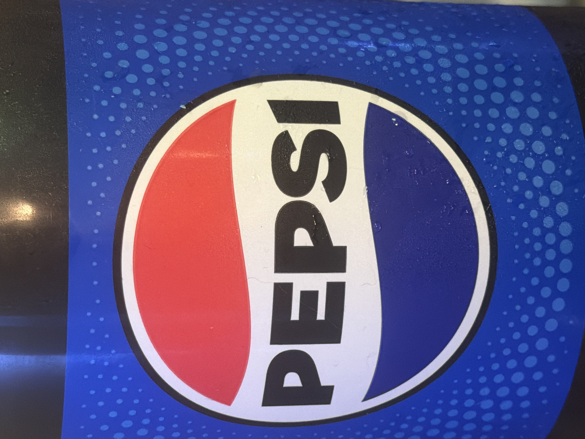 The image is a close-up shot of a Pepsi bottle, likely taken at an indoor location. The focus is on the iconic Pepsi logo, which is a red, white, and blue circle with the word "PEPSI" printed in black, bold, sans-serif letters across the center. The blue background of the bottle features a pattern of lighter blue dots of varying sizes, creating a textured effect. There are some water droplets visible on the surface of the bottle, suggesting it is cold. The top of the bottle, which appears to be black, is visible at the top of the frame. There are no people or any other identifiable objects or environmental details present in the image that would indicate a specific location within Portland, United States of America.