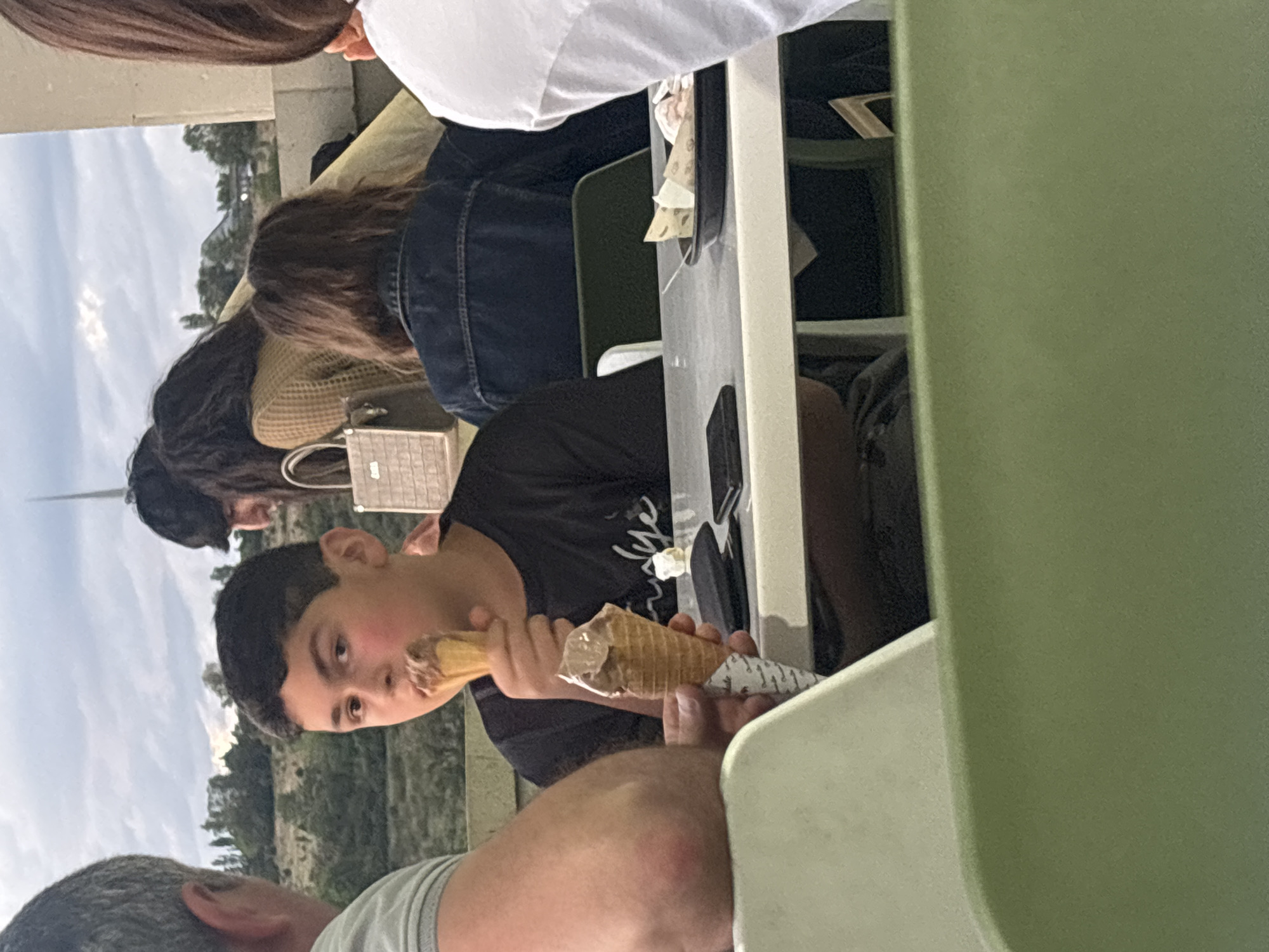 The image, oriented vertically, captures a casual gathering of several people at an outdoor or semi-outdoor dining area in Yerevan, Armenia.
In the foreground, a young boy with short, dark hair and rosy cheeks is prominently featured, looking towards the viewer. He is wearing a dark t-shirt and holding an ice cream cone wrapped in paper, which he is actively eating. To the bottom left, the partial head and shoulder of a man with short, greying hair are visible, suggesting he is seated nearby.
Further in the background, two women are seated at a white table. One woman, seen from behind, has long, dark hair and is wearing a white top. Next to her, another woman with long, dark hair is wearing a brown or tan jacket over a dark top, with a denim jacket on her back. A light-colored, patterned handbag rests beside her on the table.
The setting includes a white table and what appear to be green chairs or partitions on the right side of the frame. In the distant background, an outdoor landscape unfolds under an overcast sky, revealing trees and hills or mountains, consistent with the location context of Yerevan, Armenia.
The activity centers around the group enjoying a casual meal or snack, with the boy focused on his ice cream. The time of day appears to be daytime, possibly late afternoon, given the soft, diffused light from the cloudy sky. The overall atmosphere seems relaxed and informal. No discernible text is visible in the image.
