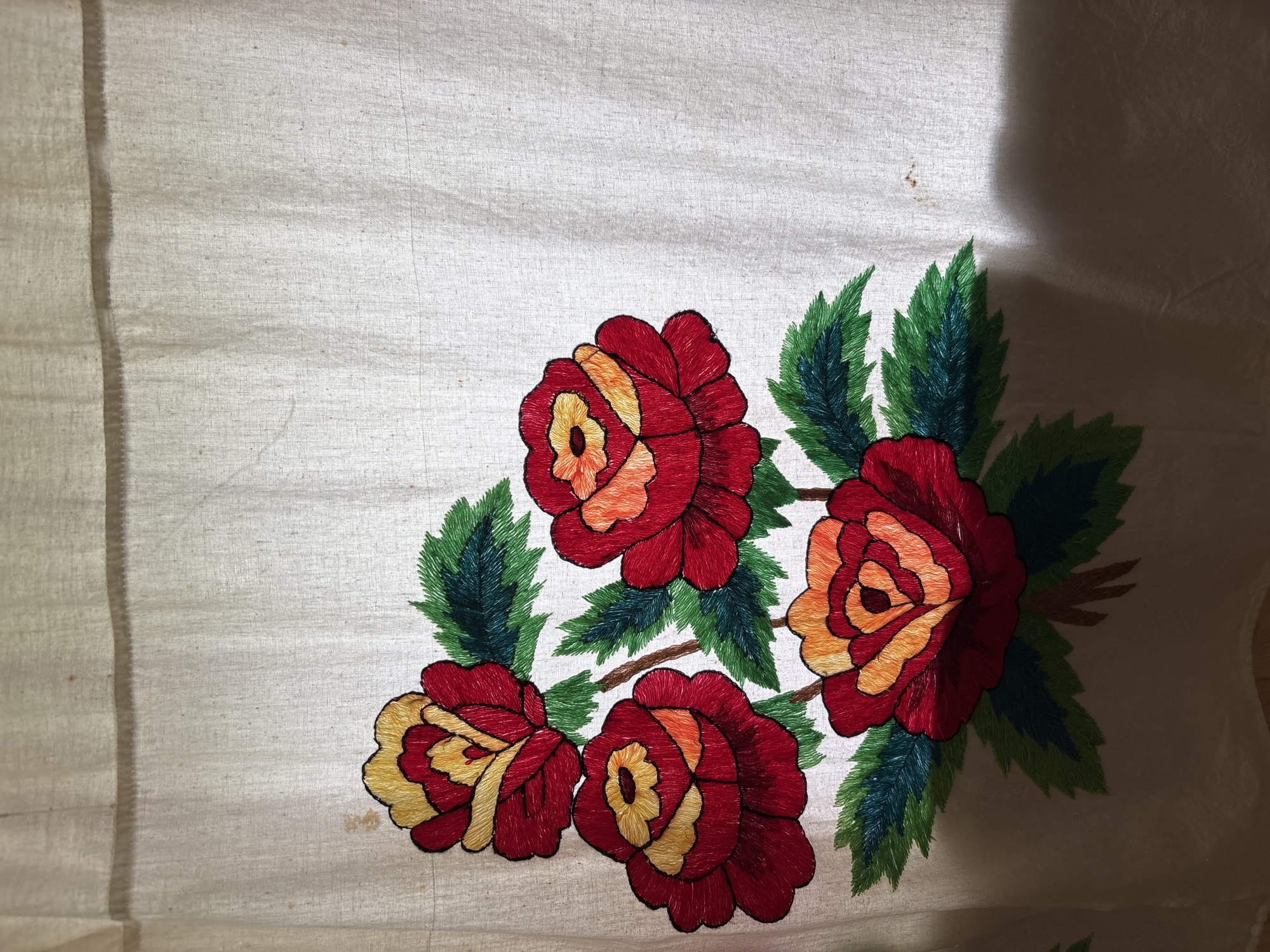 An embroidered floral pattern of four red and yellow roses with green leaves is depicted on a light-colored fabric. The embroidery is detailed, with visible stitches forming the petals and leaves. The fabric appears to be a natural fiber, possibly linen or cotton, with a slightly textured weave. The roses are arranged in a cluster, with stems and leaves extending outwards. The overall impression is of a handcrafted textile, likely a curtain or tablecloth. There are no people or any other objects visible in the image. The background is a plain, light-colored surface, possibly a wall or another piece of fabric. The image has a soft, diffused lighting. The location in the city of Ильпанур, Russia, is not discernible from the image content.
