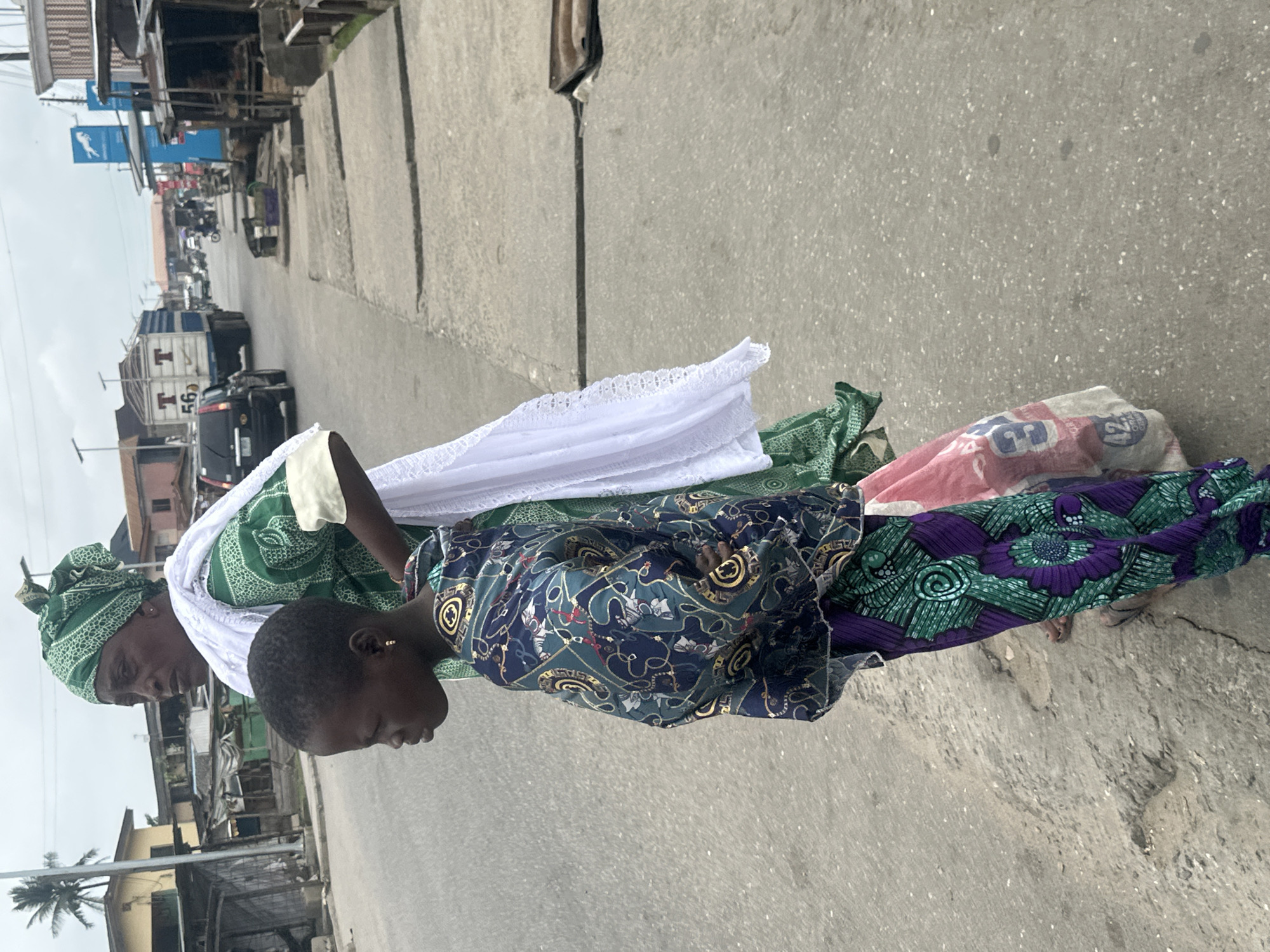 The image depicts a street scene in Badagry, Nigeria. Two individuals are standing on a cracked concrete sidewalk next to a paved road. The person in the foreground, a young child with dark skin, is facing left and wearing a vibrant, patterned long-sleeved shirt and a purple and green patterned wrap skirt. They have an earring in their left ear and their arms are crossed. Behind the child, an adult, likely a woman, is also facing left. She has dark skin and is wearing a green patterned headscarf, a green and white patterned dress, and a white shawl draped over her shoulders. A pink and white bag with blue lettering and the number "42" is visible near the adult's legs. The background shows a busy street with various buildings, including small shops with open fronts, and a large truck with "T 56" on its side driving on the road. Other vehicles and a motorcycle are also present in the distance. The sky is overcast.