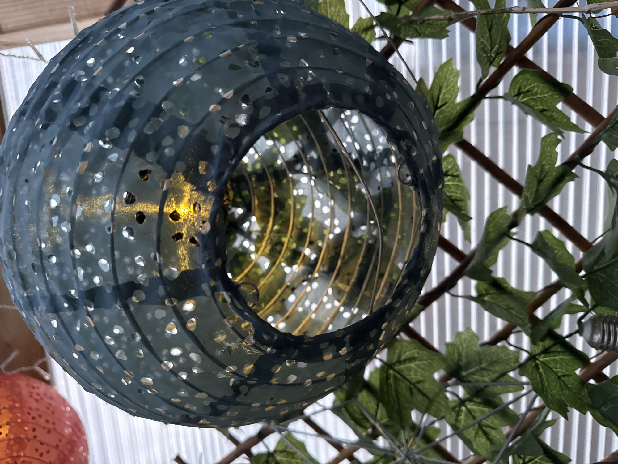 A close-up shot of a round, blue-grey paper lantern with a hole in the center and small, punched-out circles decorating its surface. The lantern appears to be lit from within, emitting a soft, golden glow. It hangs against a backdrop of a brown lattice adorned with green, artificial leaves and a white, vertically-striped surface. Another, partially visible orange lantern can be seen in the lower-left corner.