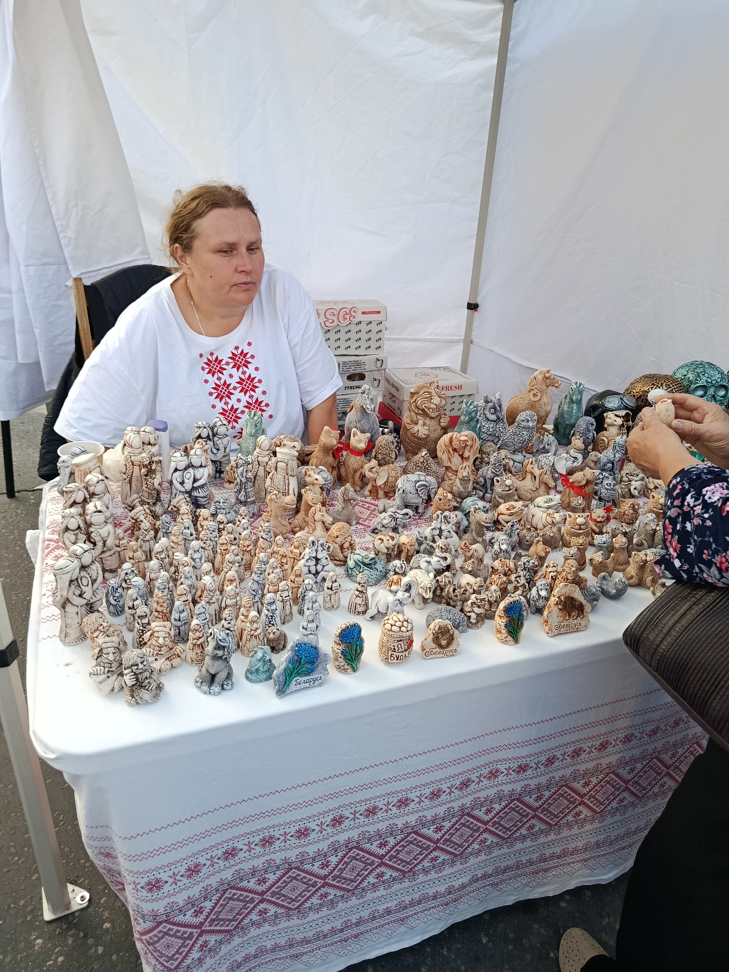 In Svietlahorsk, Belarus, a woman sells ceramic figurines at an outdoor market. She sits behind a table covered with a white tablecloth adorned with red and pink embroidered patterns. The table is laden with a vast array of small, intricately detailed ceramic figurines, primarily depicting animals and human-like figures. Among the collection are numerous small animal figurines, some wearing red ribbons. In the background, white tent fabric provides shade. A customer's hands are visible on the right side of the image, examining one of the figurines. The scene suggests a commercial transaction at a local craft fair or market in the city.