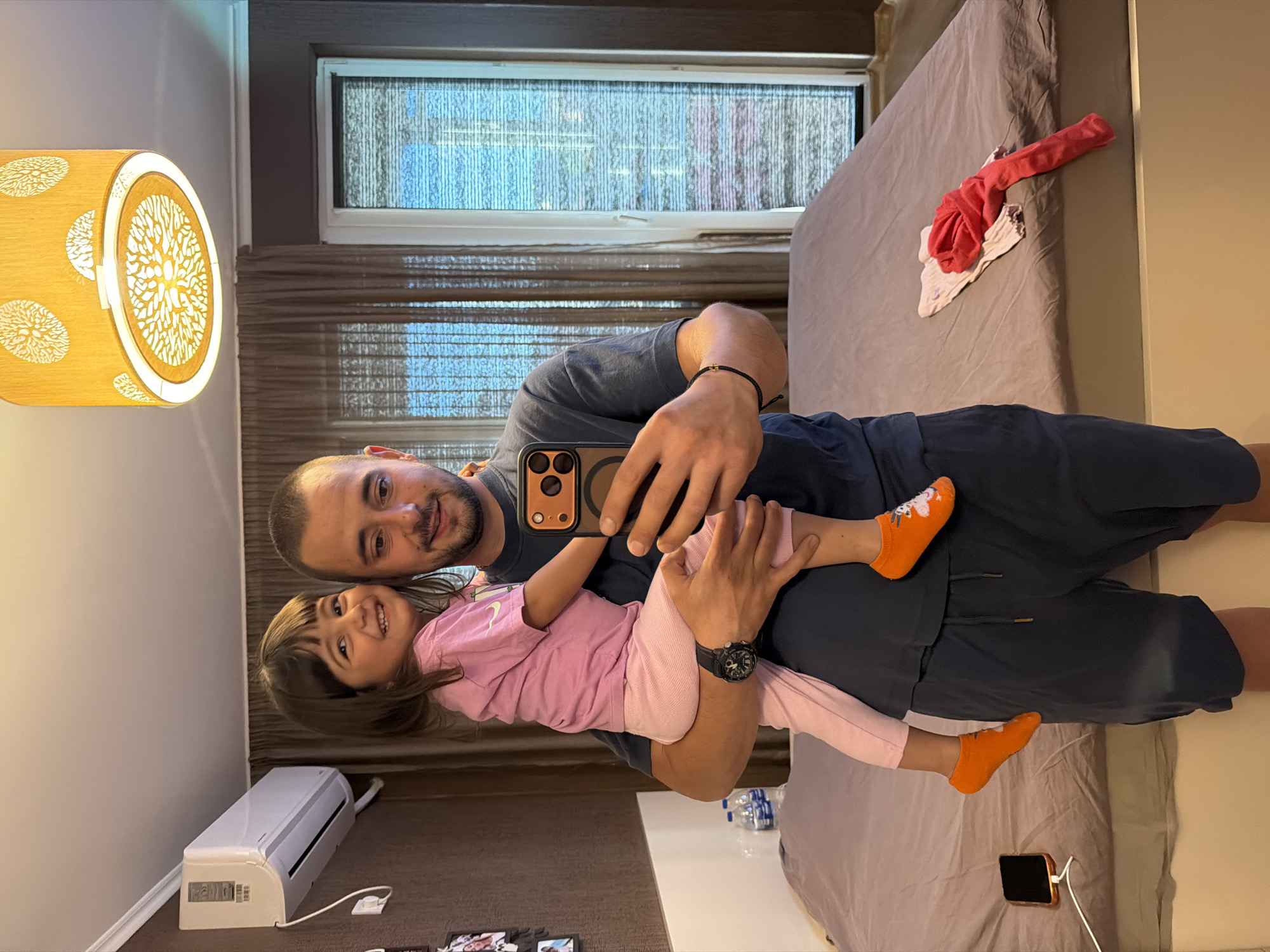 This media file is a selfie taken in a bedroom. The subjects are a man and a young girl. The man is holding the girl, who is wrapped around him. Both are smiling and appear happy. The man is wearing a dark blue t-shirt and shorts, and the girl is wearing a pink t-shirt and light pink leggings. They are both wearing bright orange socks.
In the background, a bed is visible, with some crumpled clothing on it. A window with blinds is also present. To the left of the frame, a decorative lamp with a light-colored shade and an intricate pattern is illuminated. On the floor below the window, a white air conditioning unit is mounted. There are some framed photos on the floor near the air conditioning unit. A water bottle and a mobile phone charging on a wireless charger are also visible.
The lighting suggests it might be daytime, given the light coming from the window and the lamp. The overall scene depicts a warm and intimate moment between a father and daughter in a domestic setting, likely a bedroom. The location is identified as Burgas, Bulgaria.