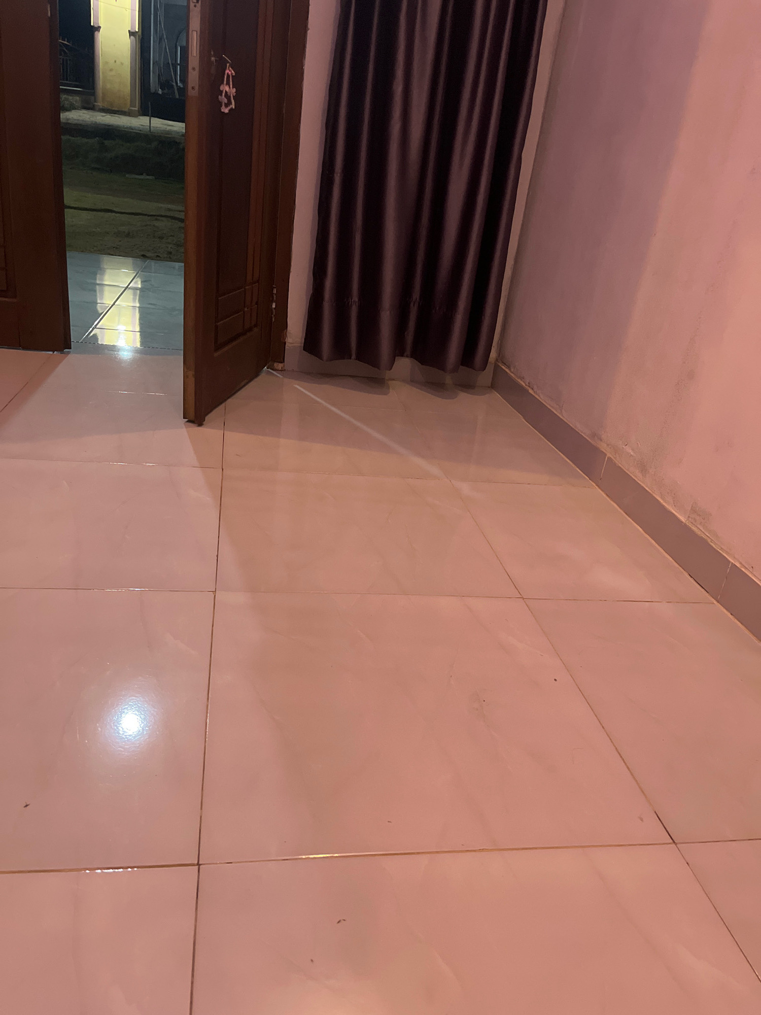 The image shows an interior view of a room with tiled flooring and a partially open wooden door. The door is on the left side of the frame, revealing a glimpse of the outdoors. A dark-colored curtain hangs on the right side of the door, covering a wall. The lighting in the room casts a pinkish hue. The floor is made of large, light-colored tiles, some of which reflect the ambient light. The scene appears to be a residential or commercial space within Kebumen, Indonesia, possibly a hallway or entryway. There are no people or specific actions depicted.