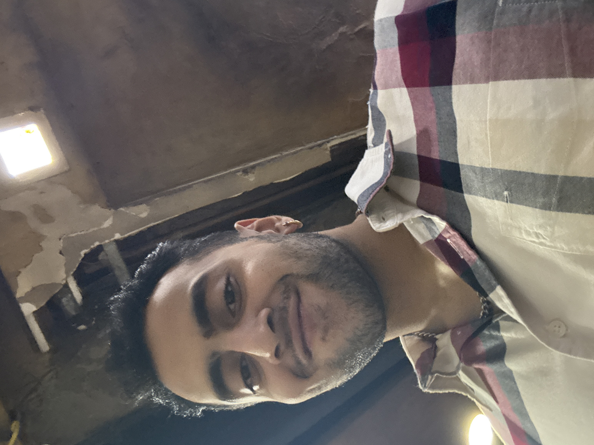 The image shows a man with dark hair and a beard wearing a checkered shirt. He appears to be indoors, with a ceiling visible in the background. A light fixture is seen on the ceiling. The photo's perspective is from below, looking up at the man. The location is unknown.
