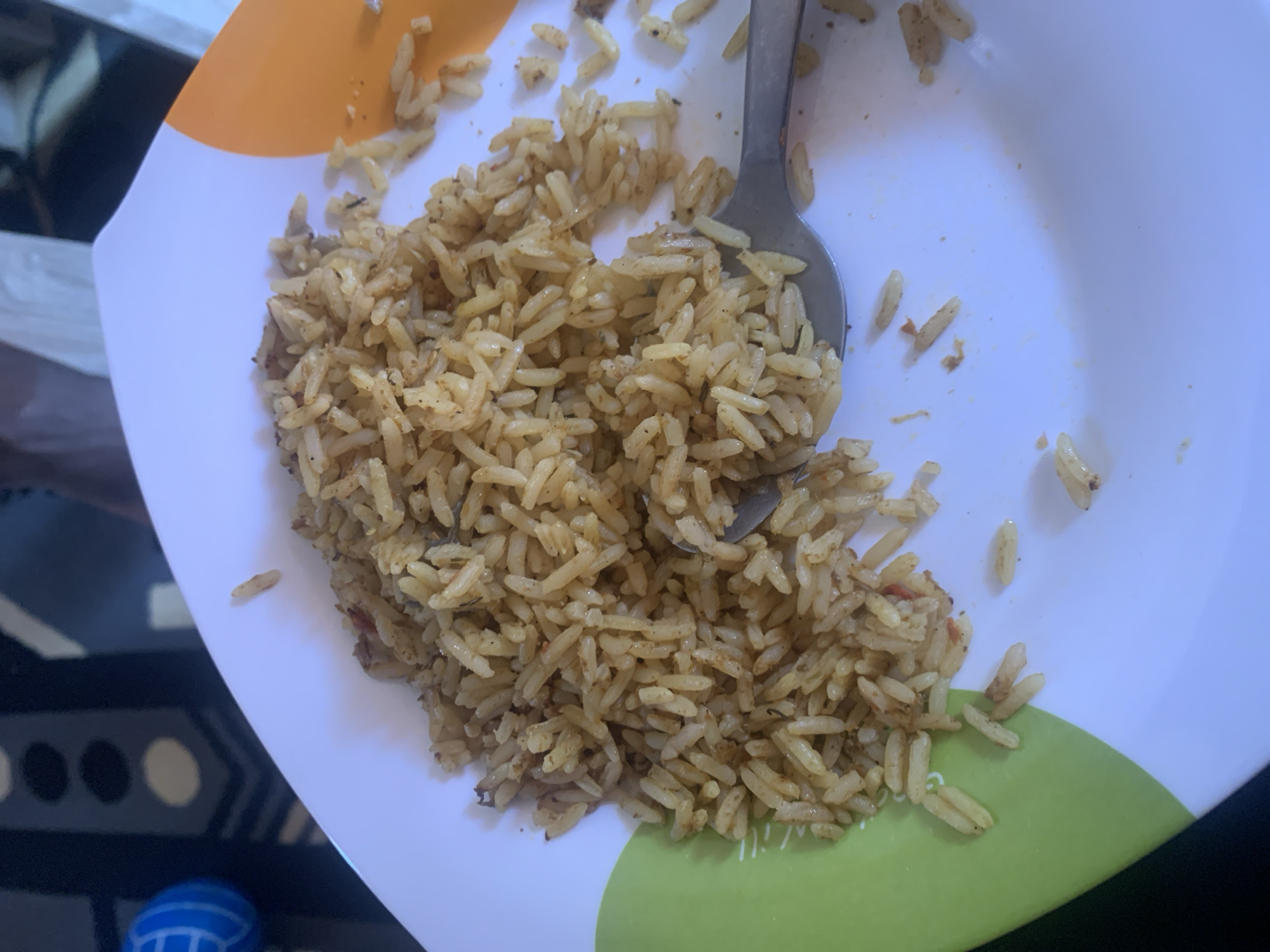 This is a close-up, overhead shot of a plate of cooked rice. The rice appears to be basmati or jasmine, with a golden-brown hue, suggesting it may be seasoned or fried. A silver spoon is partially submerged in the rice, with some grains clinging to its surface. The plate itself is white with a colorful rim that transitions from orange to green. Scattered grains of rice are visible outside the main pile, on the surface of the plate. The background is dark and out of focus, with some hints of what could be furniture or a tablecloth. The lighting suggests an indoor setting, possibly during the day. There are no people visible, and the focus is entirely on the food. The overall impression is of a simple meal about to be eaten. The location context "Gura Top, Nigeria" is not directly evident from the visual cues in the image.
