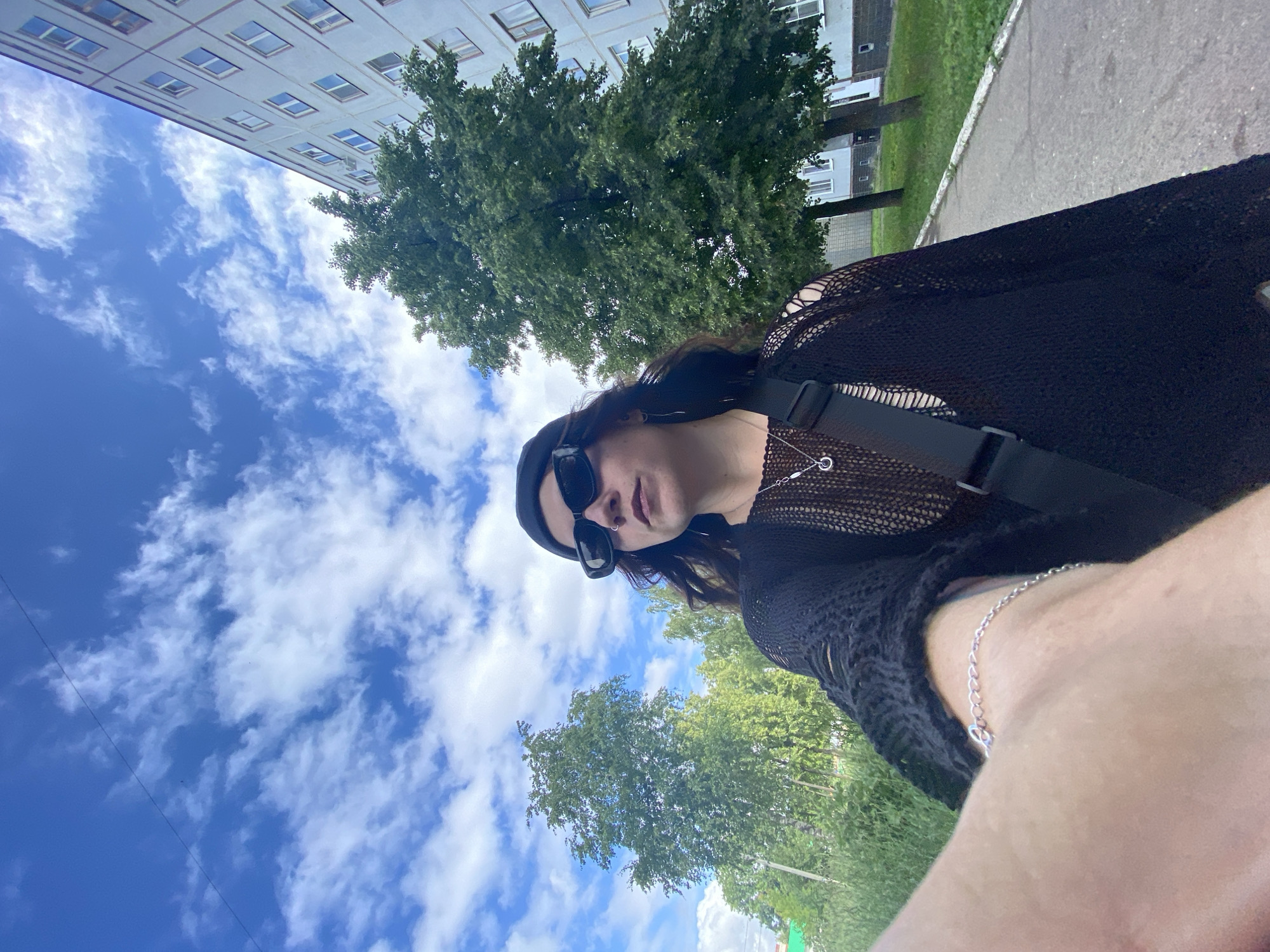 A person, wearing black sunglasses, a black beanie, and a black mesh sweater over a tank top, is taking a selfie outdoors. They are wearing a silver necklace and a silver bracelet. They have a black bag with a black strap slung across their chest. The person is standing on a paved walkway next to a grassy area with trees. A light blue apartment building is visible in the background. The sky is blue with white clouds. The location is Chaikovsky, Russia.