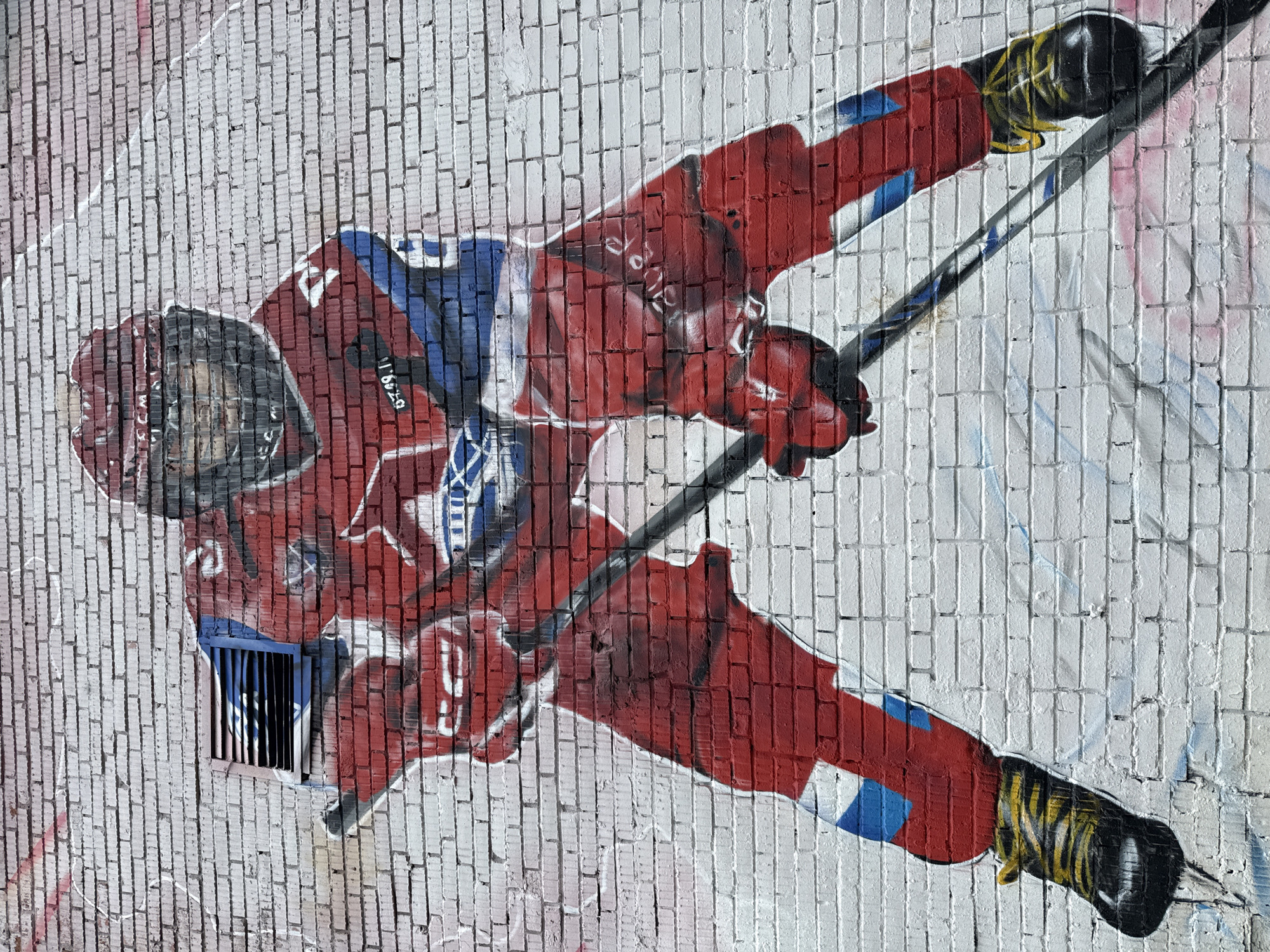 A graffiti mural depicts a hockey player in a red and blue uniform, skating with a hockey stick. The mural is painted on a white brick wall. The player is wearing a red helmet and visor, and red gloves. The uniform has white and blue accents. The player's skates are black with yellow laces. The mural is located in Rusanovo, Russia. The style of the graffiti is realistic, with shading and detail suggesting movement and depth. The brick texture of the wall is visible beneath the paint, adding to the raw, urban feel of the artwork. There are no other people or objects visible in the immediate vicinity of the mural. The lighting suggests an outdoor setting, possibly during the daytime.