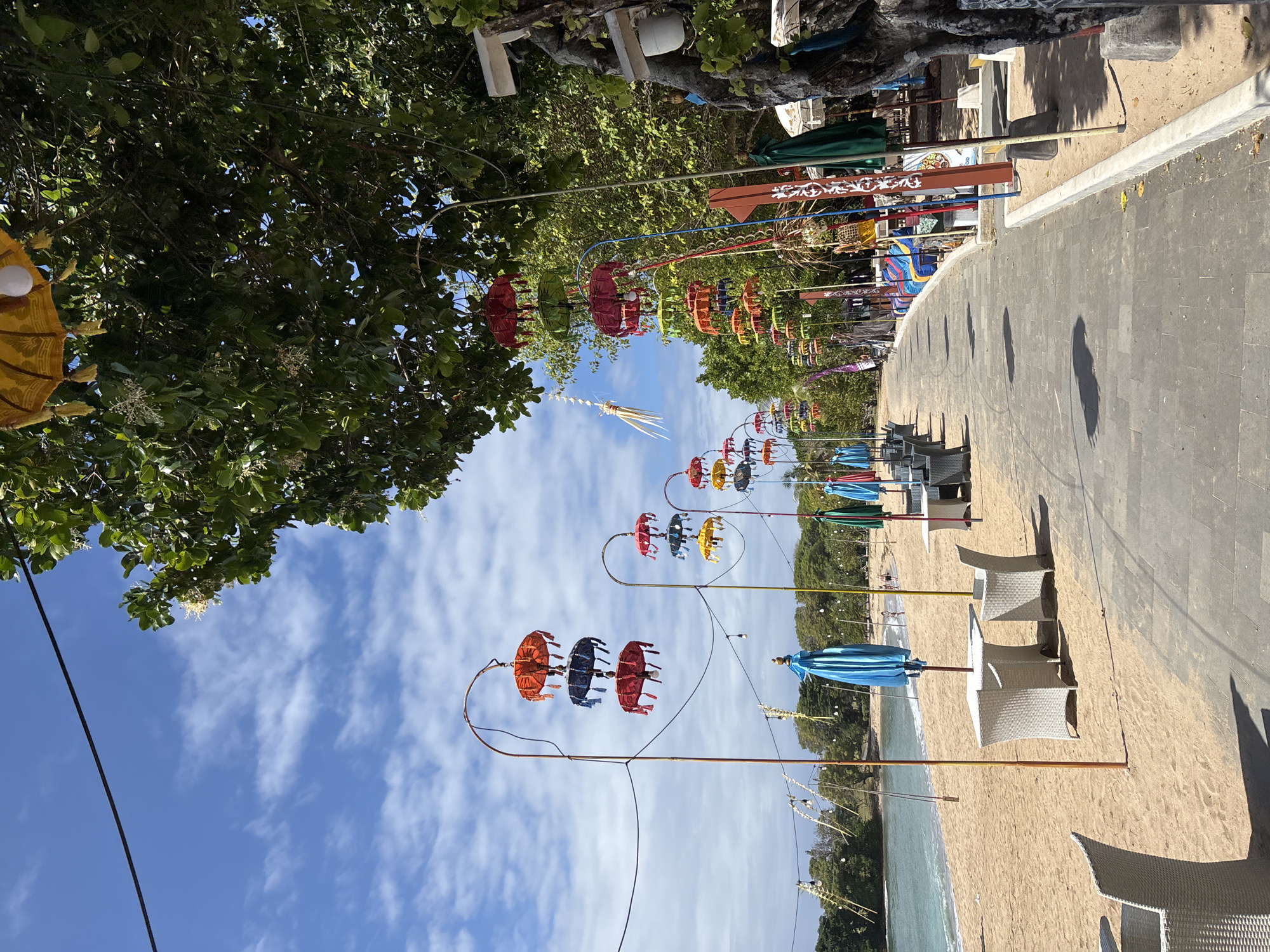 The image depicts a beachside promenade in Kuta Selatan, Indonesia, under a bright blue sky with scattered clouds. A paved walkway lines the sandy beach, which is dotted with white wicker tables and chairs, suggesting a dining or relaxation area. Along the promenade, colorful decorative umbrellas hang in clusters from tall poles, adding a vibrant touch to the scene. Trees with lush green foliage border the walkway, providing shade. The ocean is visible on the left, with gentle waves lapping the shore. A few distant figures can be seen on the beach, indicating a light presence of people. The overall impression is a serene and inviting tropical setting, characteristic of a beach resort.