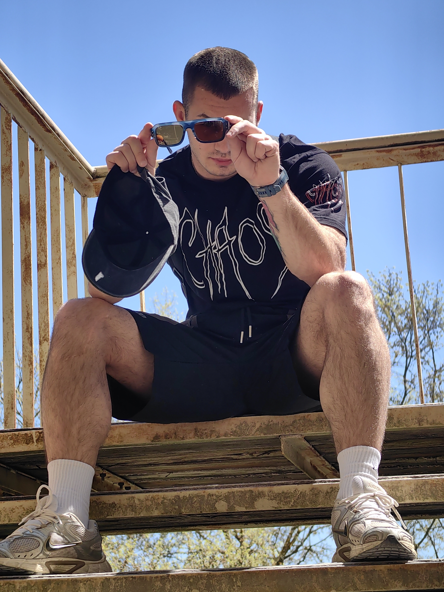 A man is sitting on a rusted metal staircase outdoors on a sunny day. He is wearing a black t-shirt with a white design, black shorts, white socks, and dirty grey athletic shoes. He is holding sunglasses in his right hand and a black baseball cap in his left hand, partially obscuring his face. He has a watch on his left wrist and tattoos on his left arm. The staircase has a rusted metal railing. Trees are visible in the background. It is not possible to definitively identify the location within Varna, Bulgaria from the image alone.