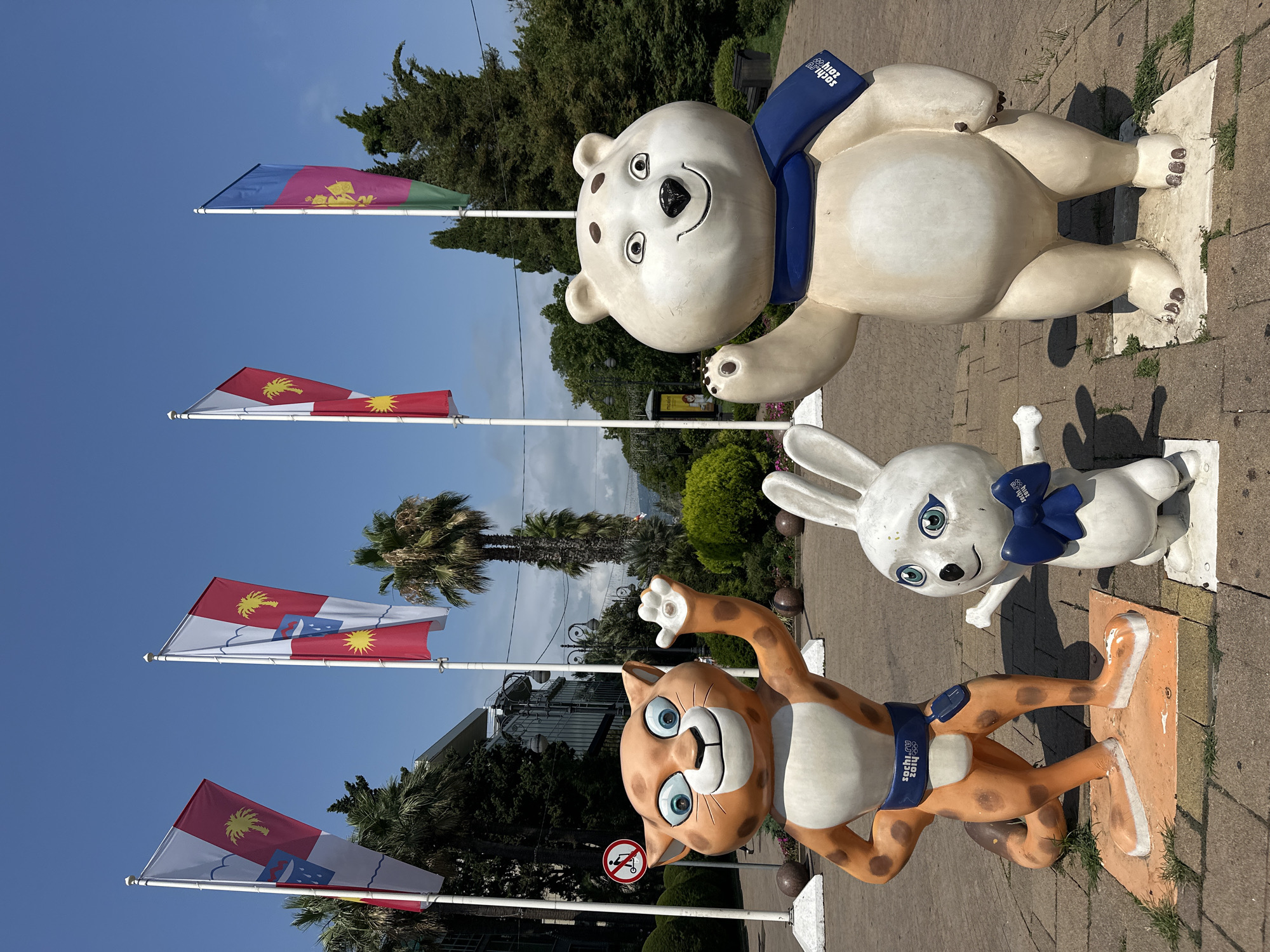 The image depicts a sunny outdoor scene featuring three mascot statues in Sochi, Russia. On the left is a leopard-like creature with orange fur and blue eyes, wearing a blue belt with "Sochi 2014" and the Olympic rings. In the center is a white rabbit with blue eyes and a blue bow tie, also bearing the "Sochi 2014" logo. To the right stands a polar bear, also white, with blue eyes, a blue scarf with the "Sochi 2014" logo, and dark brown spots on its head. The statues are positioned on a paved walkway, with a no-smoking sign visible in the background to the left. Behind the statues, several flags are raised on white poles against a clear blue sky, interspersed with palm trees and other greenery. A modern building with glass windows is visible behind the statues and flags. The overall environment suggests a public space, possibly a park or promenade in Sochi, likely related to the 2014 Winter Olympics.