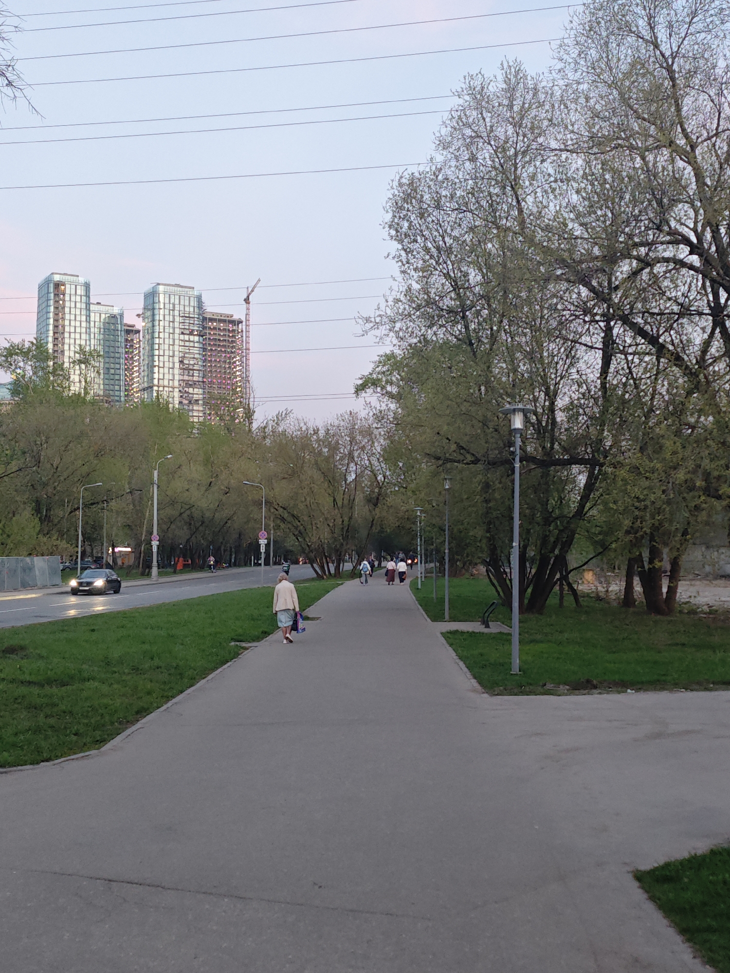 The image captures a street scene in Moscow, Russia, at dusk. In the background, tall, modern buildings and a construction crane suggest a developing urban area. A six-lane road divides the image horizontally; a black car travels along the road. A woman walks away from the camera on a wide, paved sidewalk. Several other pedestrians stroll along the path further down. Trees line both sides of the sidewalk and road, adding greenery to the urban landscape. Light posts illuminate the area. Power lines stretch across the sky, adding to the urban backdrop.