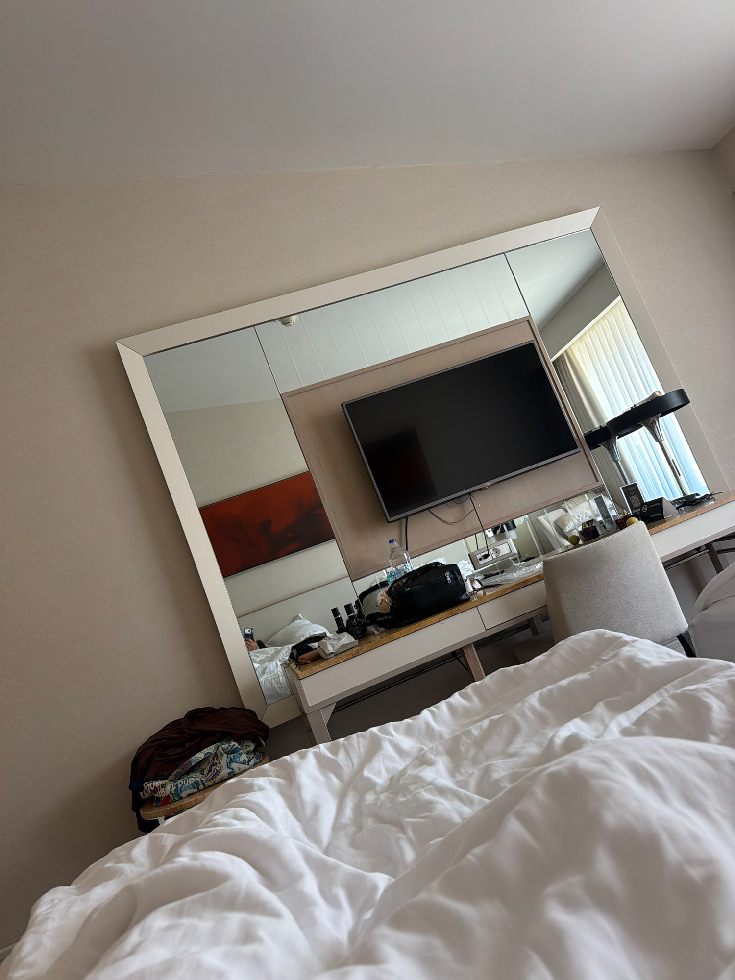 The image shows a messy hotel room in Kuşadası, Turkey. A large mirror is mounted on the wall, reflecting the room. A flat-screen television is mounted above a desk. On the desk are various items, including a bottle of water, a black bag, a chair, and toiletries. A pile of clothes is on the floor next to the desk. The bed is unmade and covered in white sheets. The overall impression is of a lived-in and somewhat cluttered space, likely a hotel room.