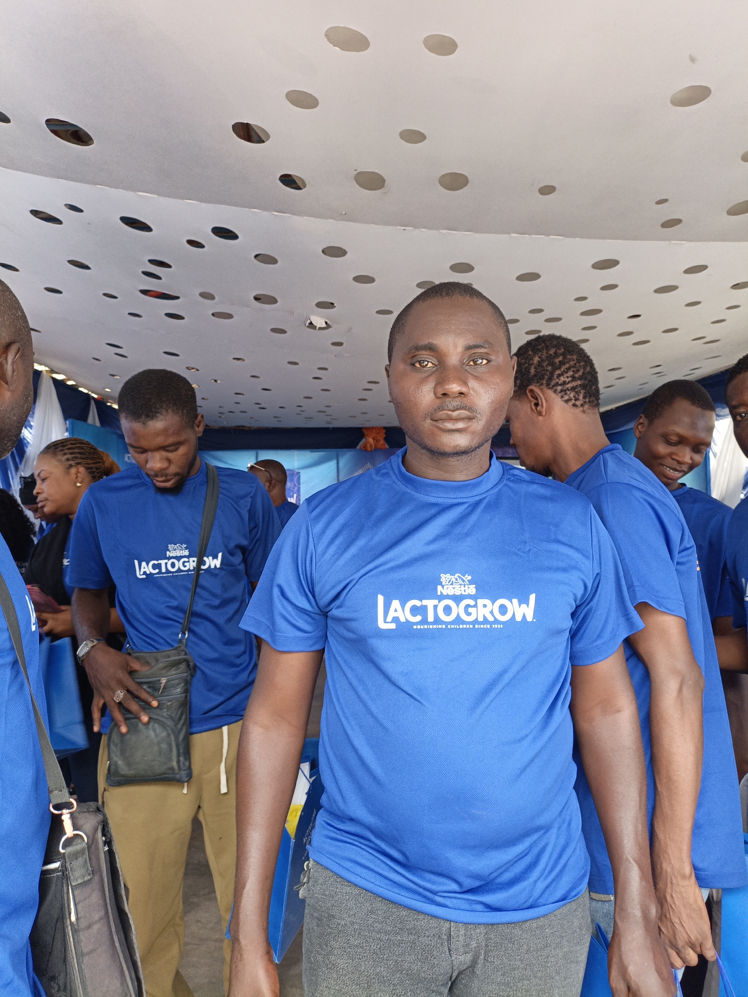 A group of predominantly men, all wearing identical bright blue t-shirts, are gathered under a white canopy with a distinctive polka-dot pattern on its underside. The setting appears to be an indoor or covered outdoor event in Tarauni, Nigeria.
The central figure, a man with a neutral expression, looks directly at the camera. His blue t-shirt clearly displays the white "Nestlé LACTOGROW" logo and the tagline "NOURISHING CHILDREN SINCE 1921." Several other men in similar attire are visible around him; one man to his right looks away, another further back smiles, and a man to his left looks down, wearing a black shoulder bag. Another man is partially visible on the far left, also in a blue shirt and carrying a larger black bag. A woman can be seen in the background, partially obscured, behind one of the men.
The matching branded t-shirts suggest that the individuals are part of an organized event, likely a promotional or staff gathering for Nestlé Lactogrow, a product focused on child nutrition. The background includes decorative blue and white striped fabric and a faint blue banner, indicating an event space. The lighting is bright, consistent with a daytime event. The scene depicts a candid moment within what appears to be a busy gathering.